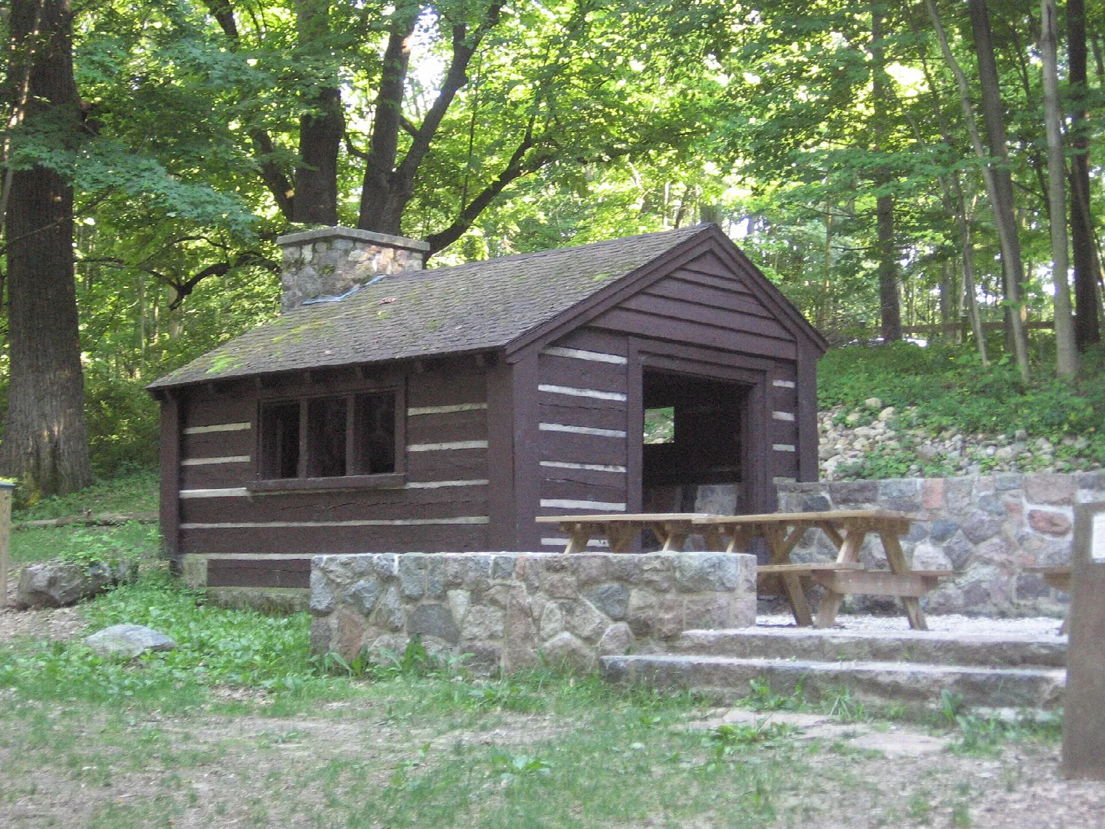Small log shelter with a stone base and steps in a green wooded area
