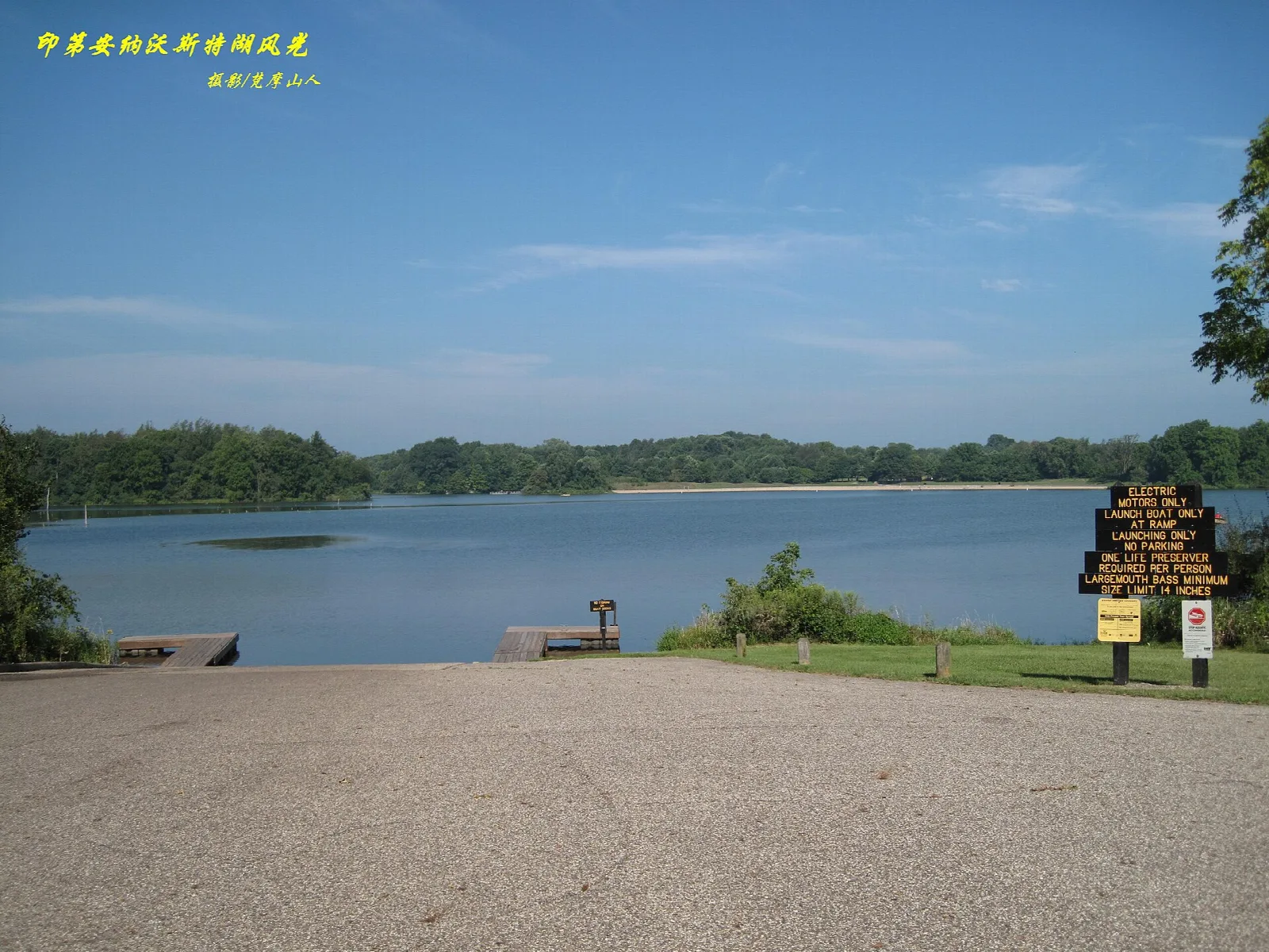 Lake with wooded shore seen from a gravel boat launch, with a sign at the right