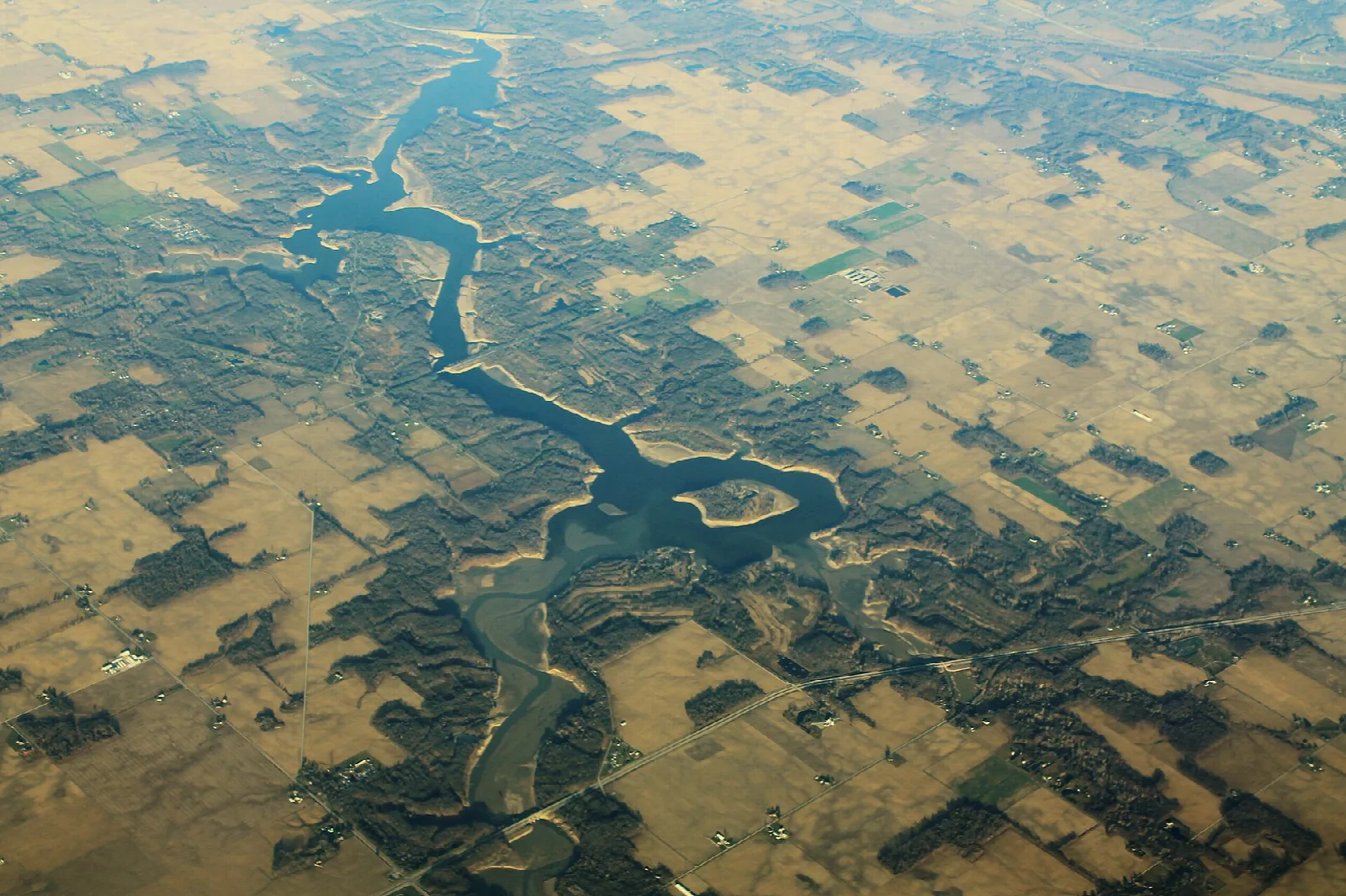 Aerial view of a winding lake and dam among tan farm fields and dark woods