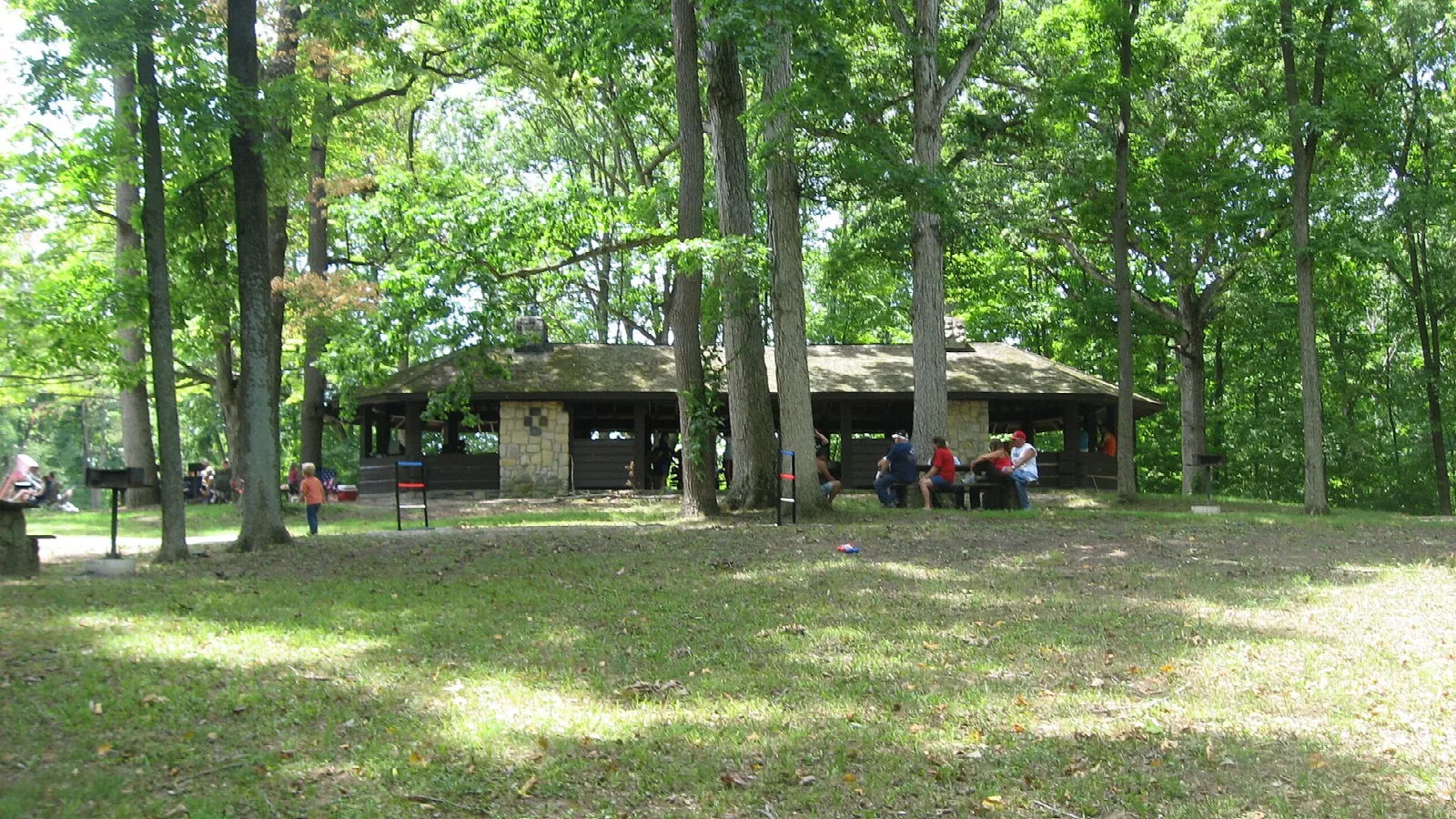 Open-sided stone and timber picnic shelter among tall trees, with people at tables