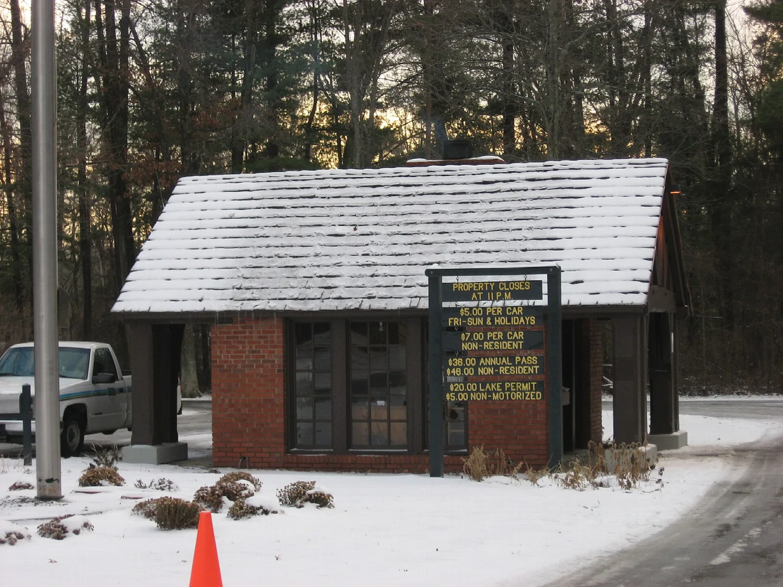 Small brick gatehouse with a snowy roof beside a road, with pine trees behind
