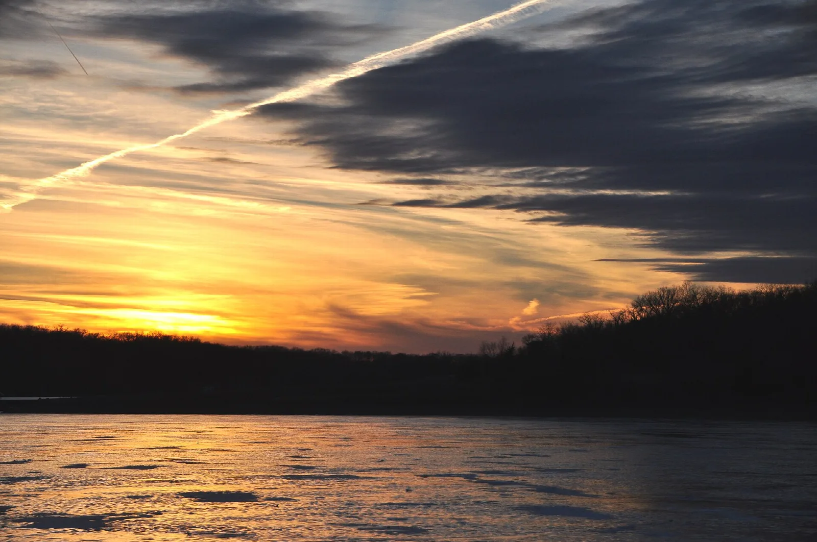 Orange sunset over dark lake water with jet trails