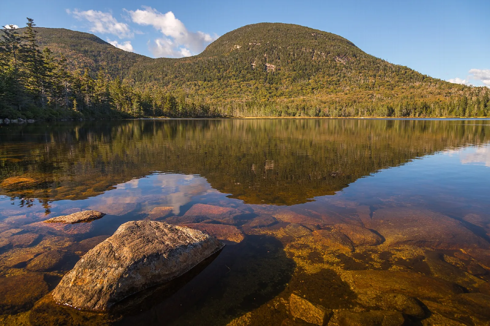 Calm lake reflecting a forested mountain, with rocks visible under clear water in the foreground