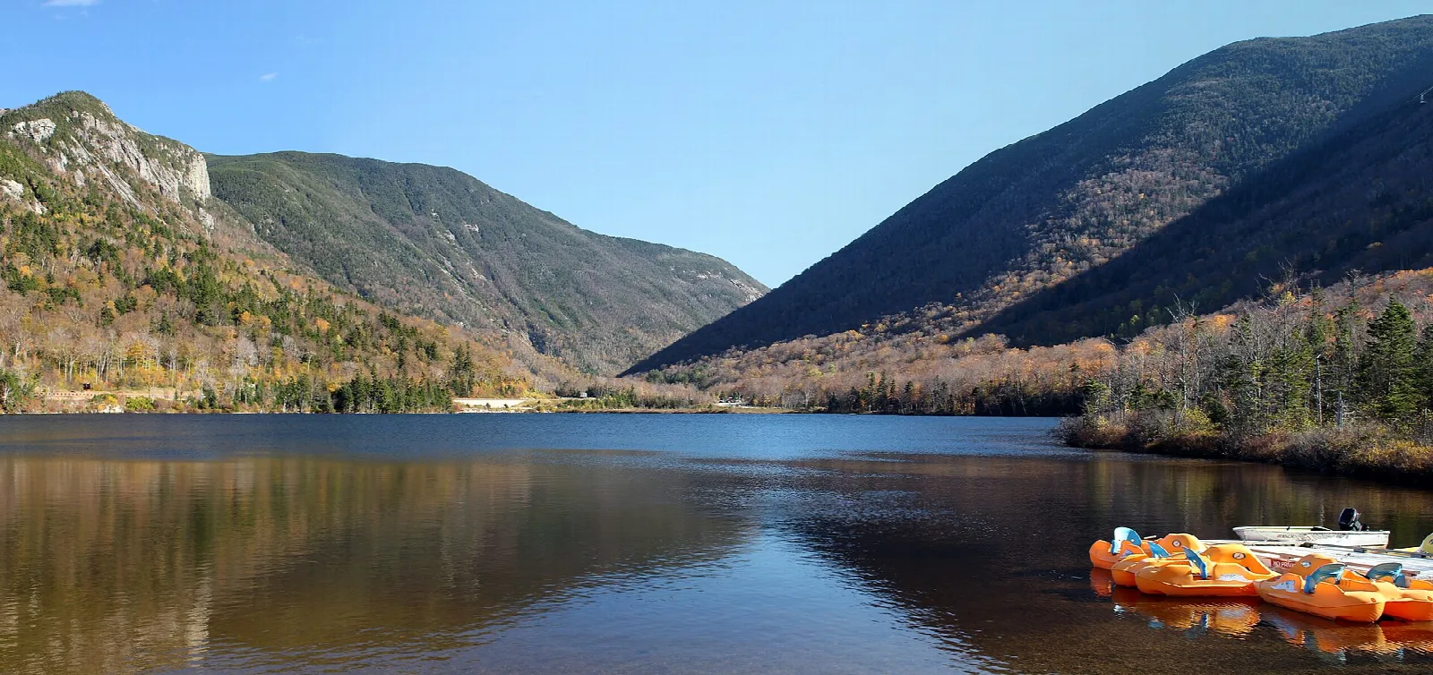 Mountain lake with forested ridges on both sides and orange boats on the right shore