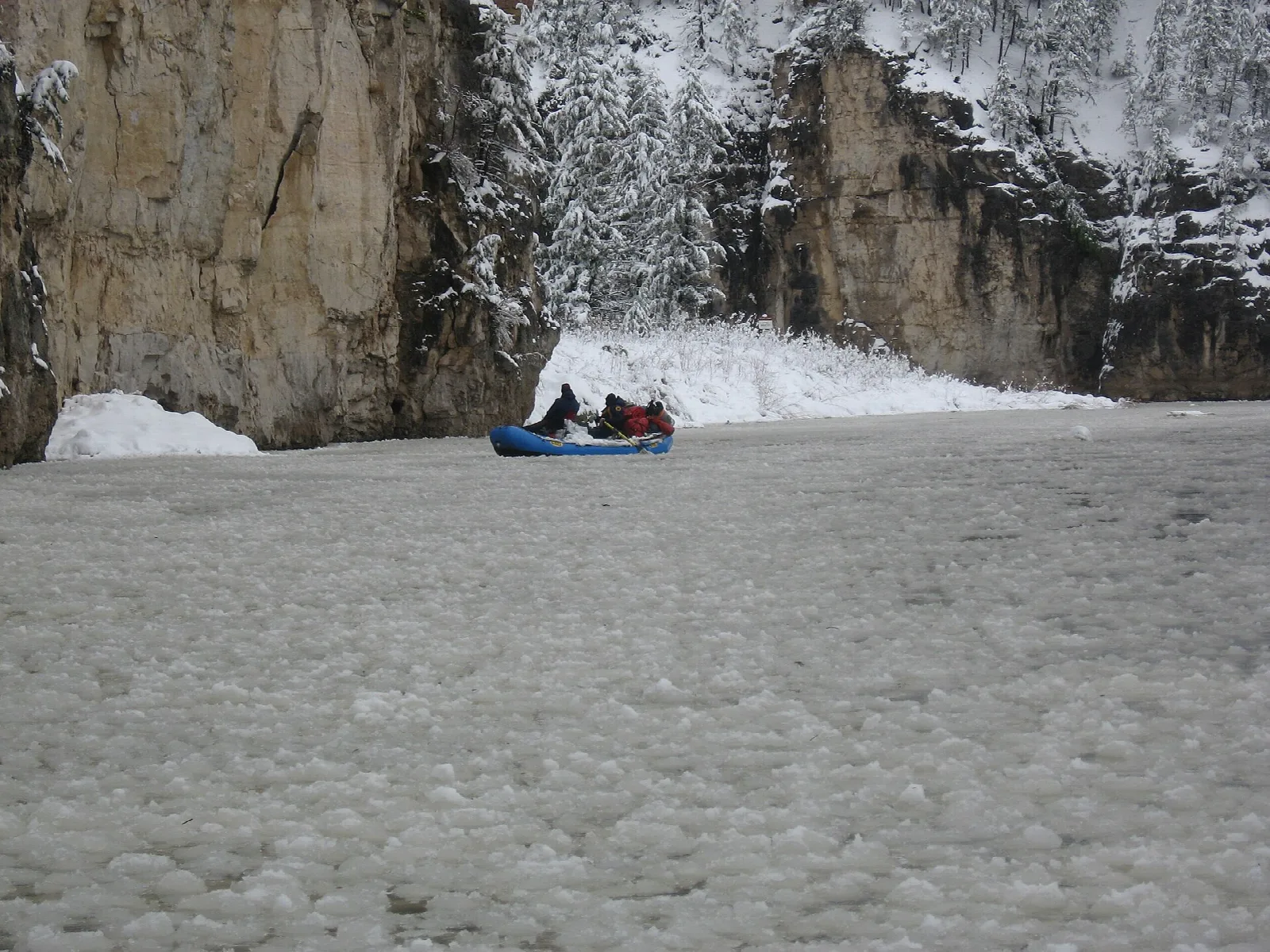 Small blue raft on an icy river below a snowy rock wall