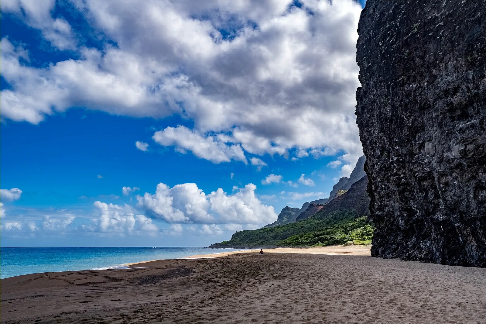 Empty sandy beach beside a tall dark cliff, with blue sky and clouds