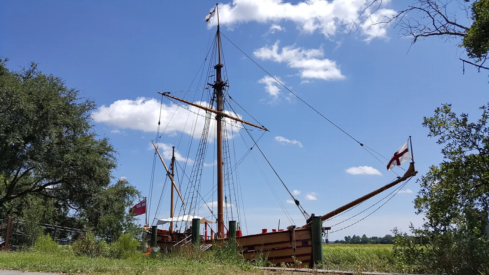 Wooden ketch with tall masts docked beside marsh grass under blue sky