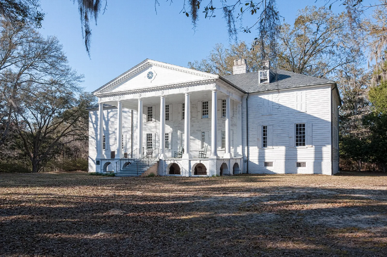 White columned house with a portico, seen from a gravel yard under bare trees