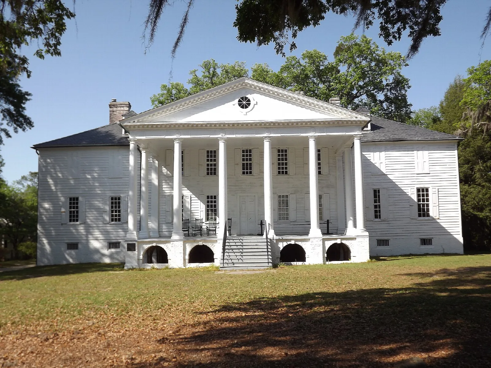 White plantation house with a tall columned portico and brick arches below, on a lawn