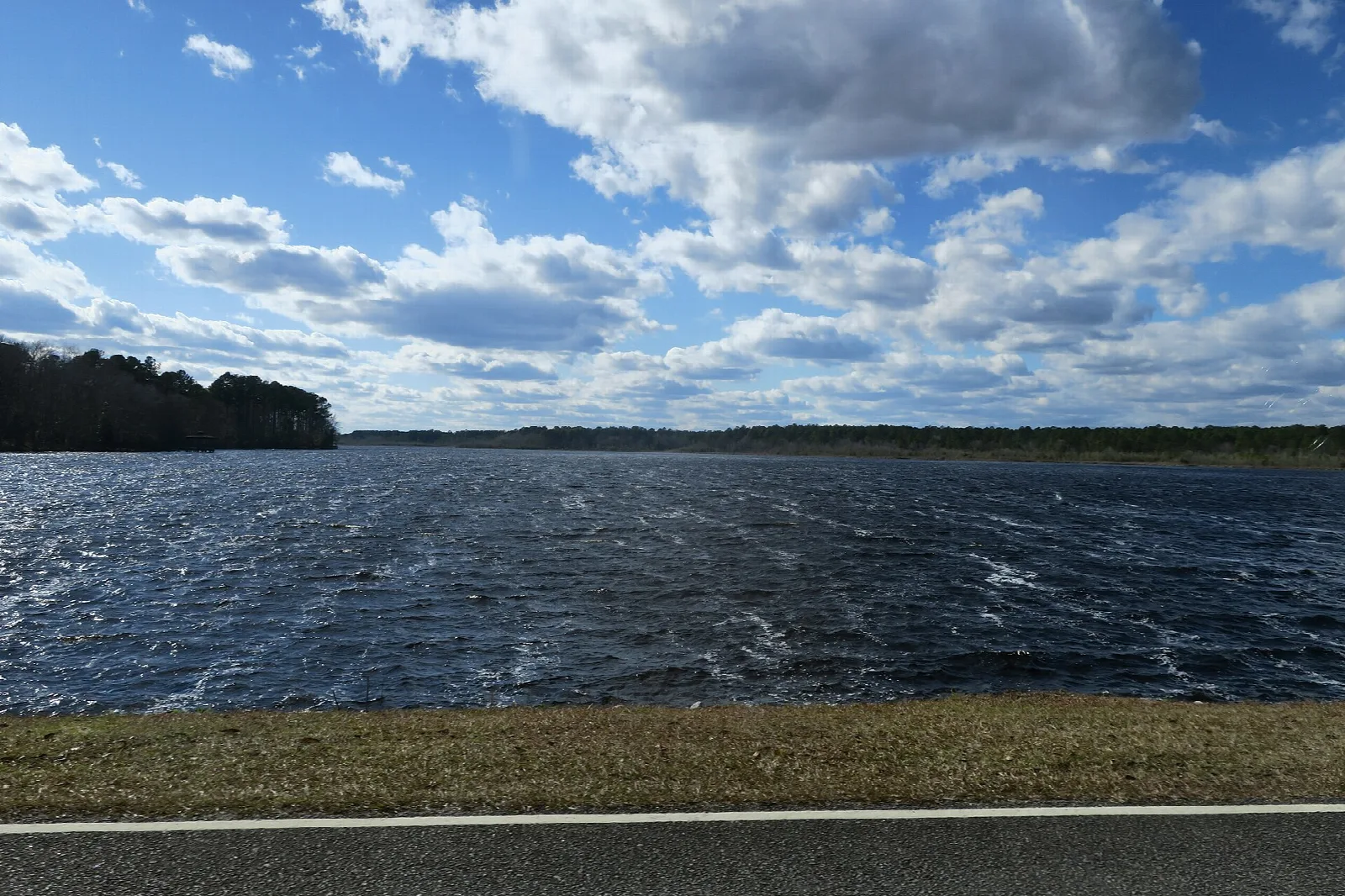 Wide lake with choppy dark water under a blue sky with clouds, seen over a road edge