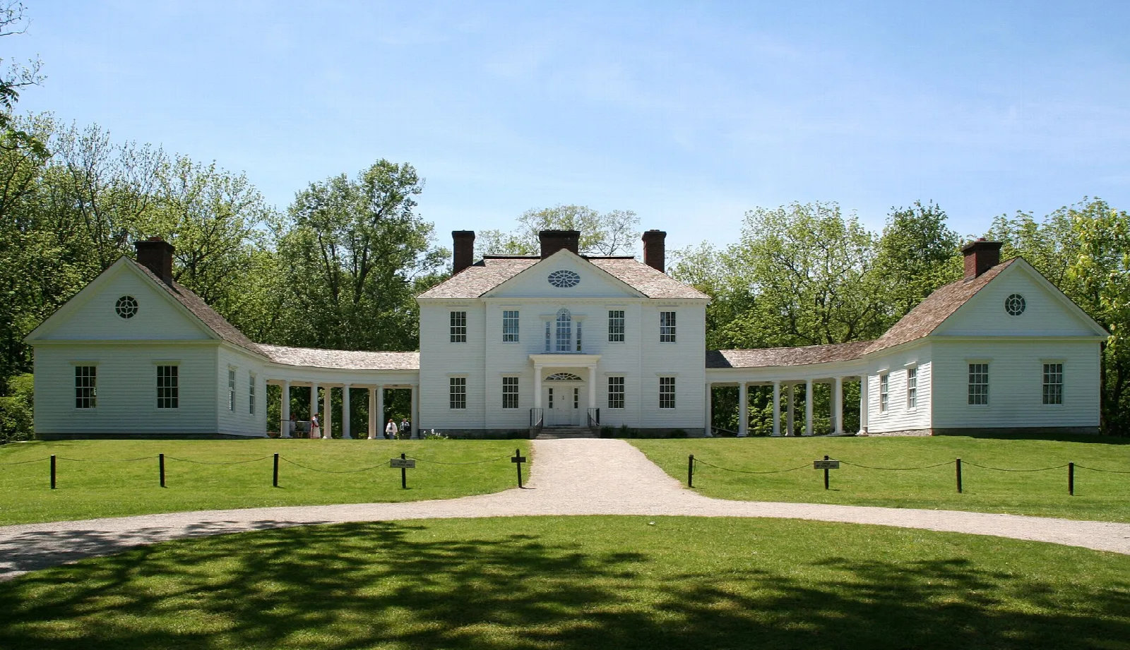 White Palladian mansion with two wings, a curved path and green lawn under blue sky