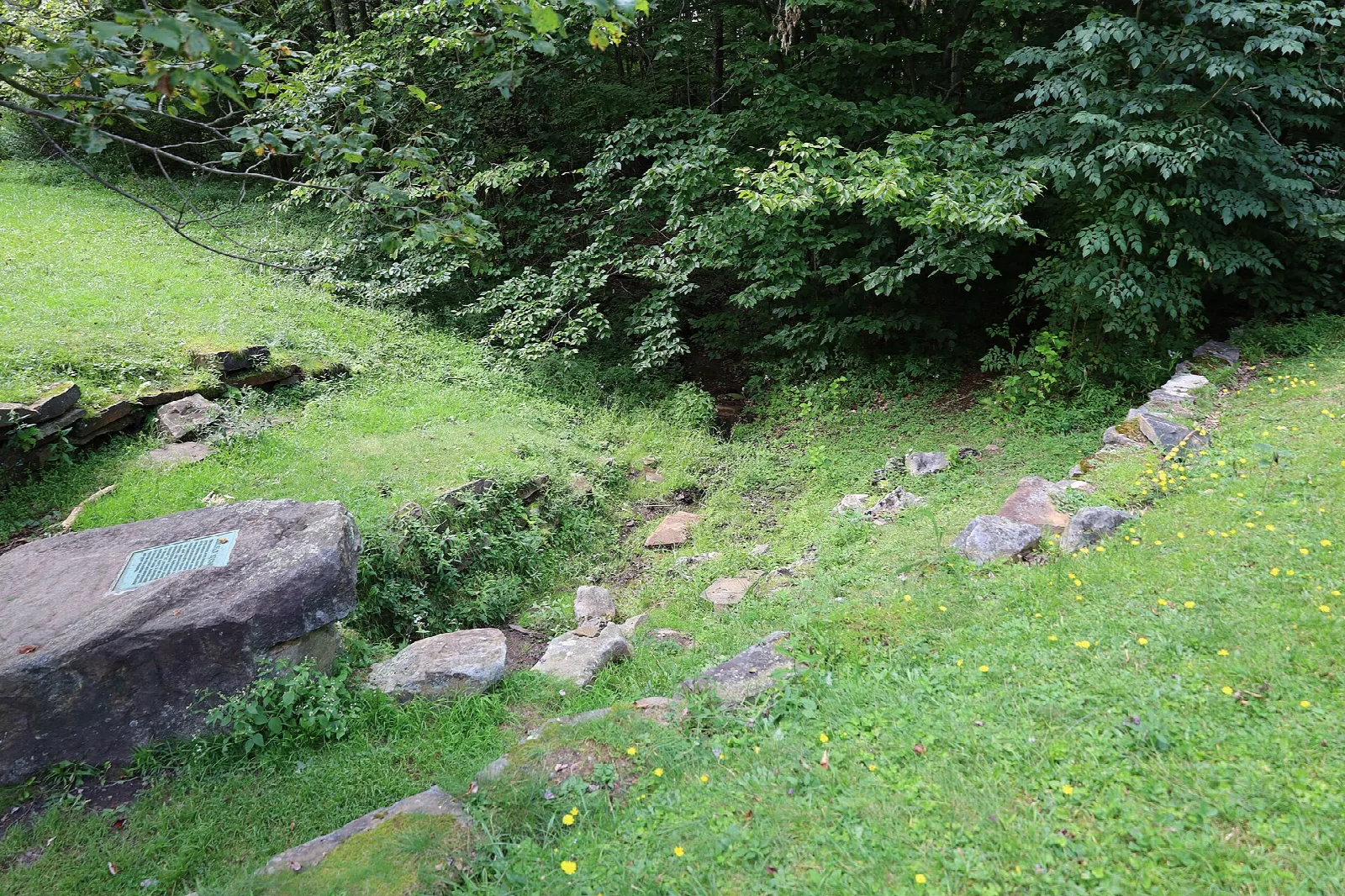 Large boulder with a plaque beside a stone wall and green woods
