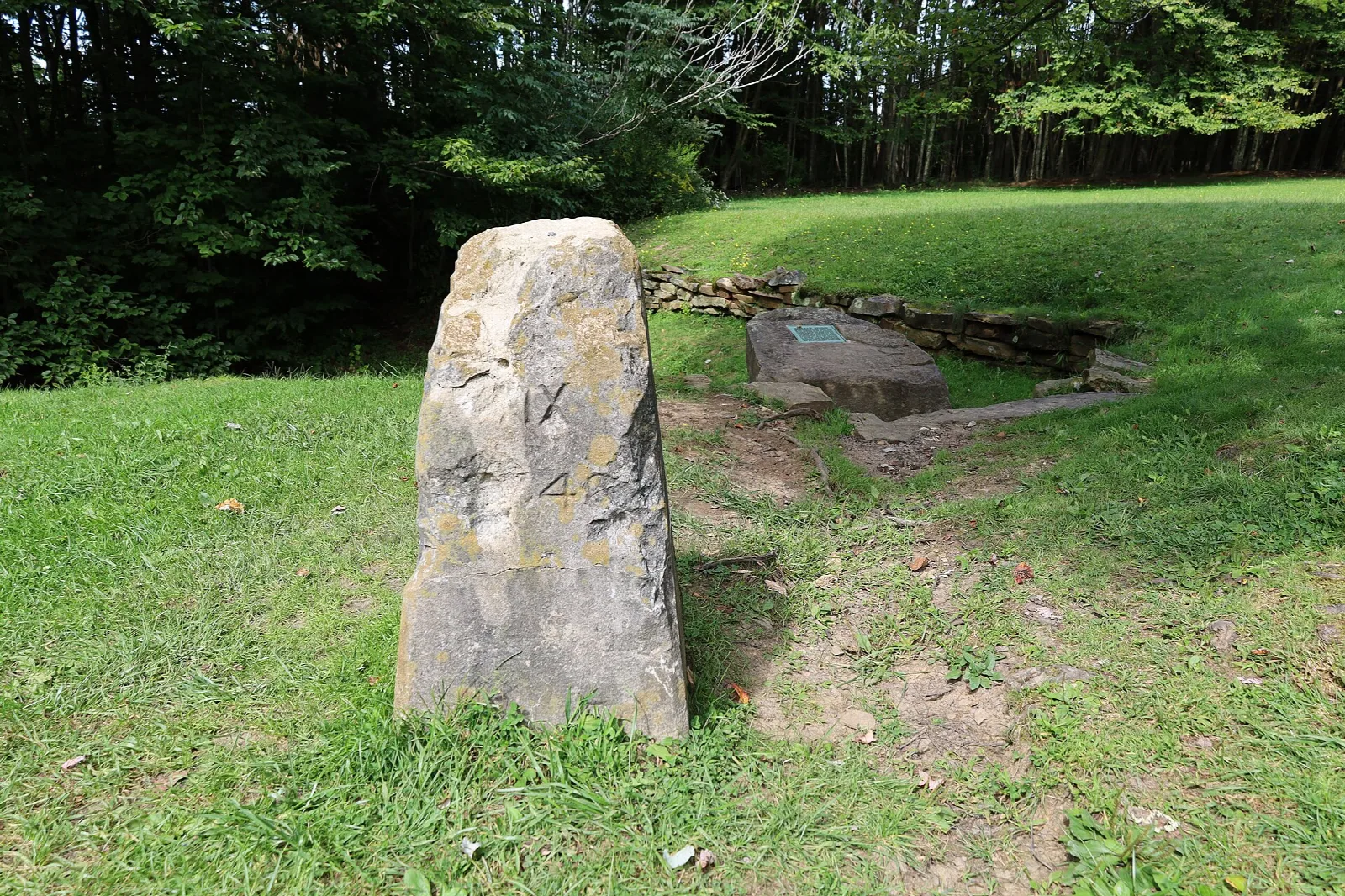 Upright stone marker on a mown lawn with a plaque stone behind it and trees beyond