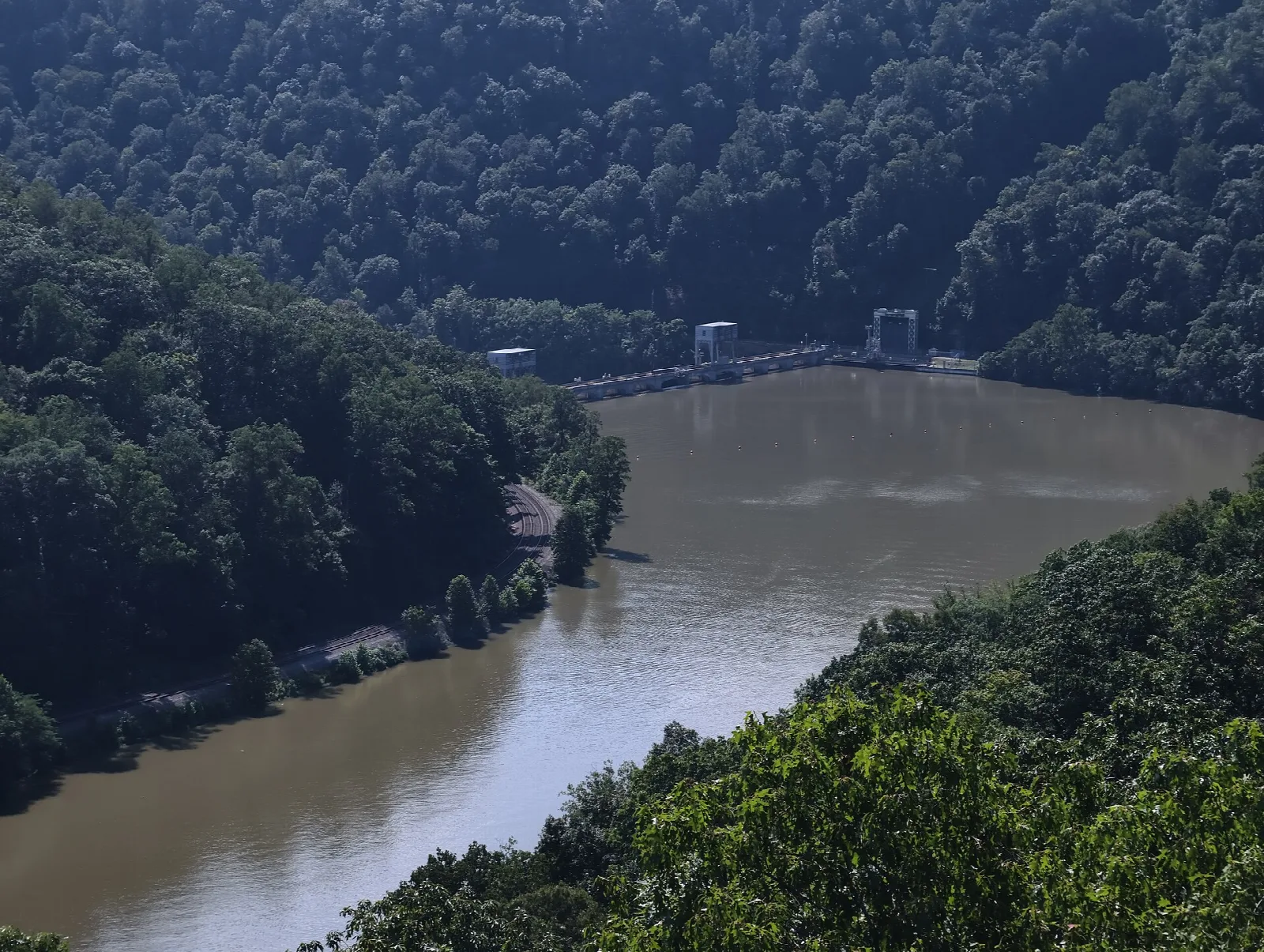 Brown river below forested hills with a small dam in the distance