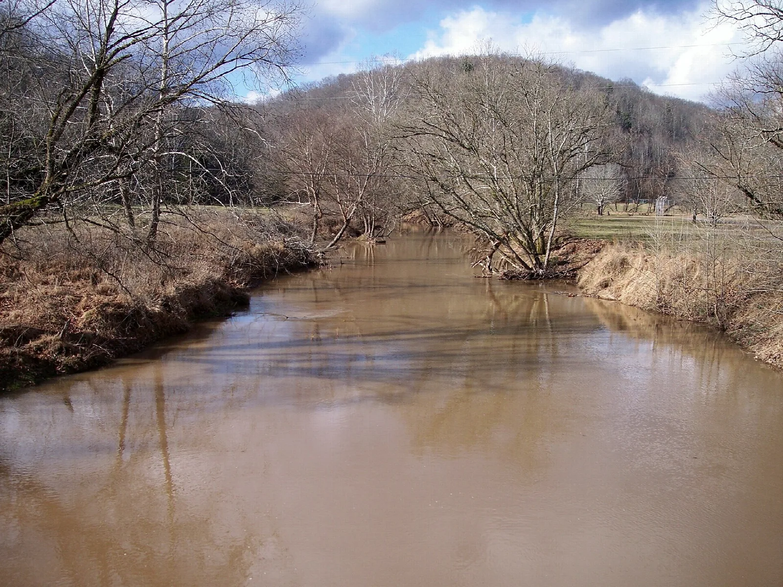 Brown creek running between bare brown banks with a wooded hill beyond
