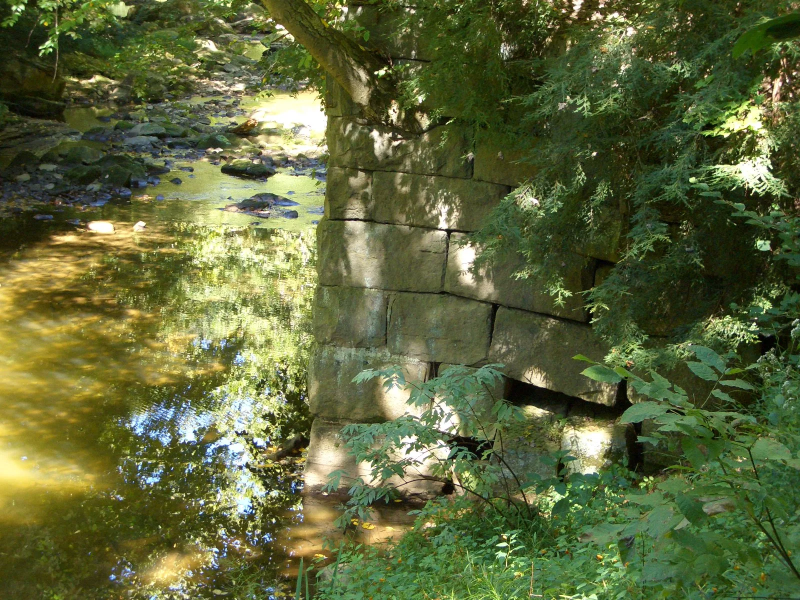 Stone abutment beside a sunlit stream under green trees