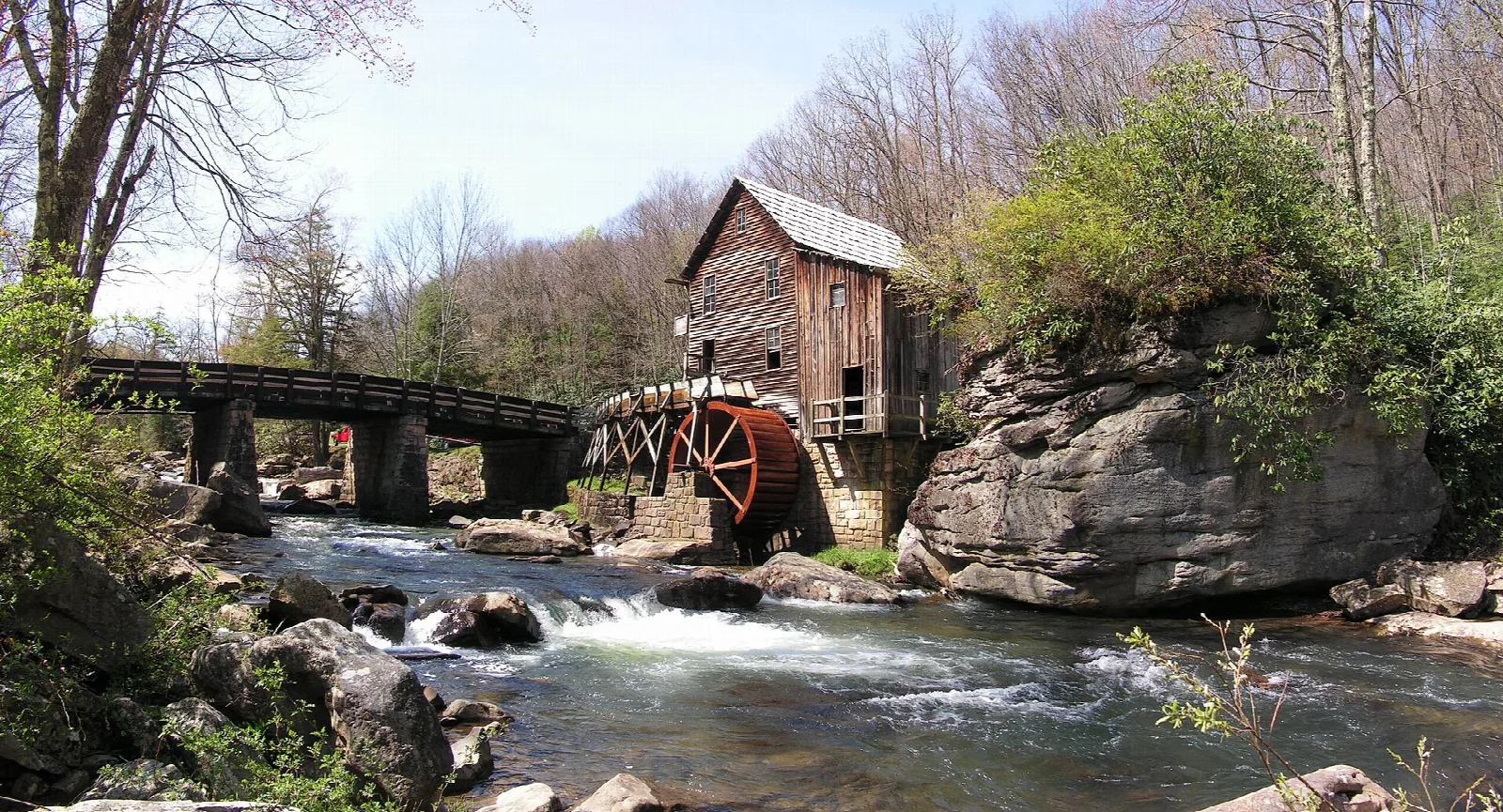 Wooden grist mill with a water wheel beside a rocky creek and footbridge