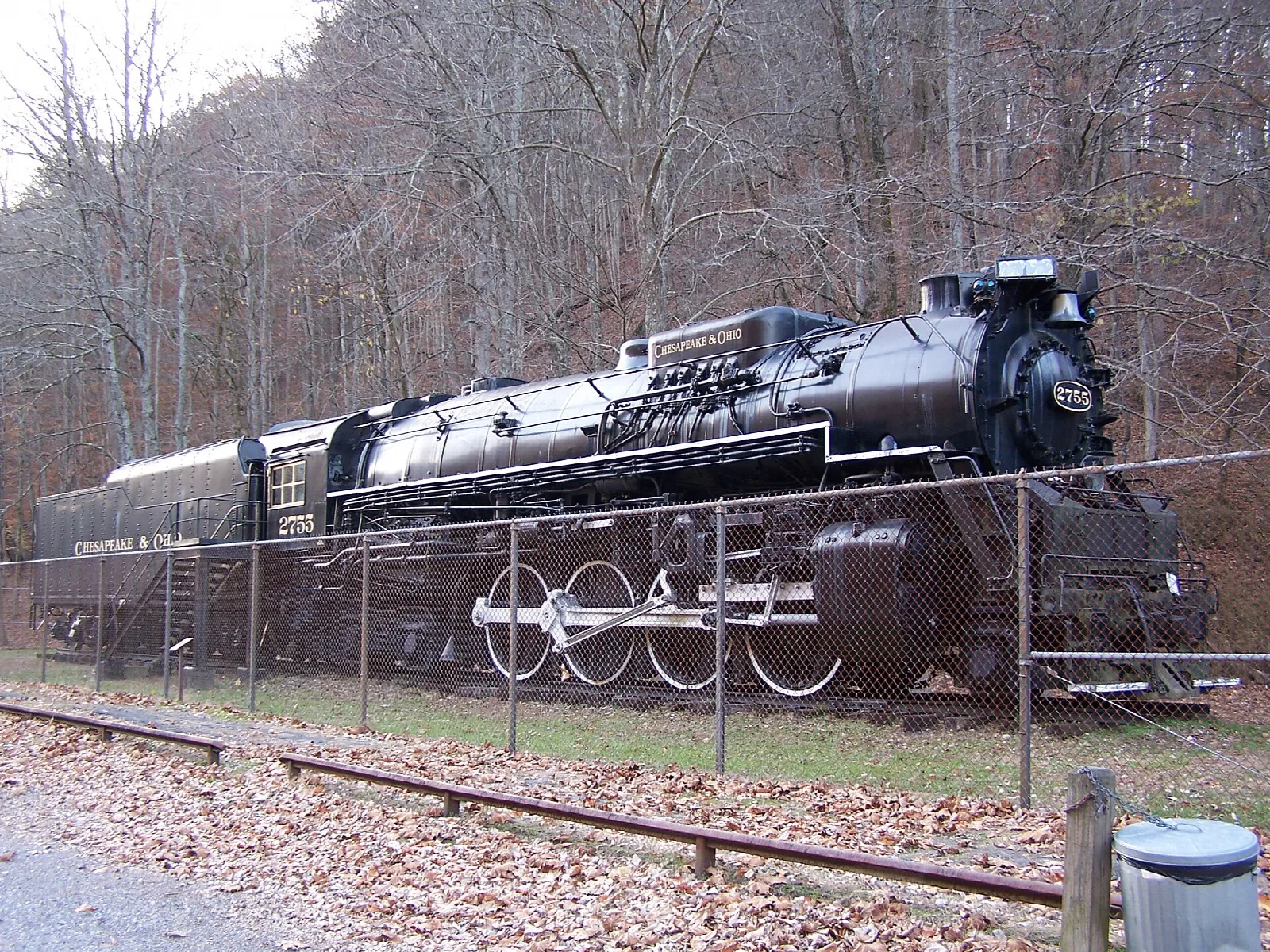 Large black steam locomotive on display behind a fence beside a wooded hillside