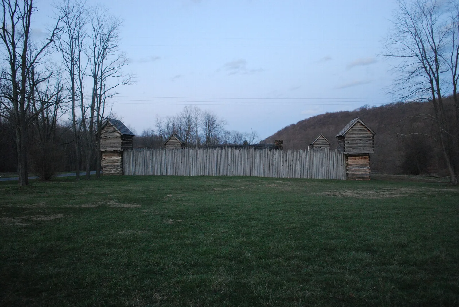Wooden stockade wall with blockhouses across a grass field at dusk