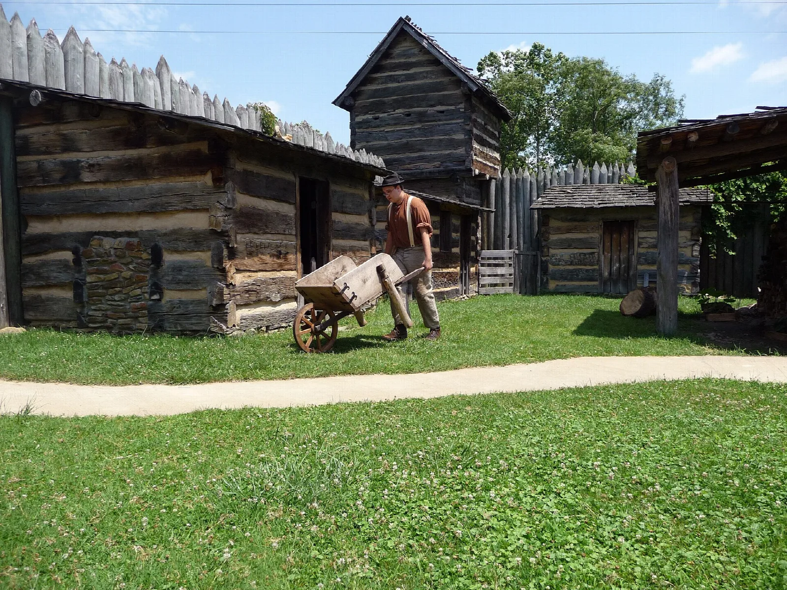 Log cabins inside a wooden stockade with a person pushing a wheelbarrow across the grass