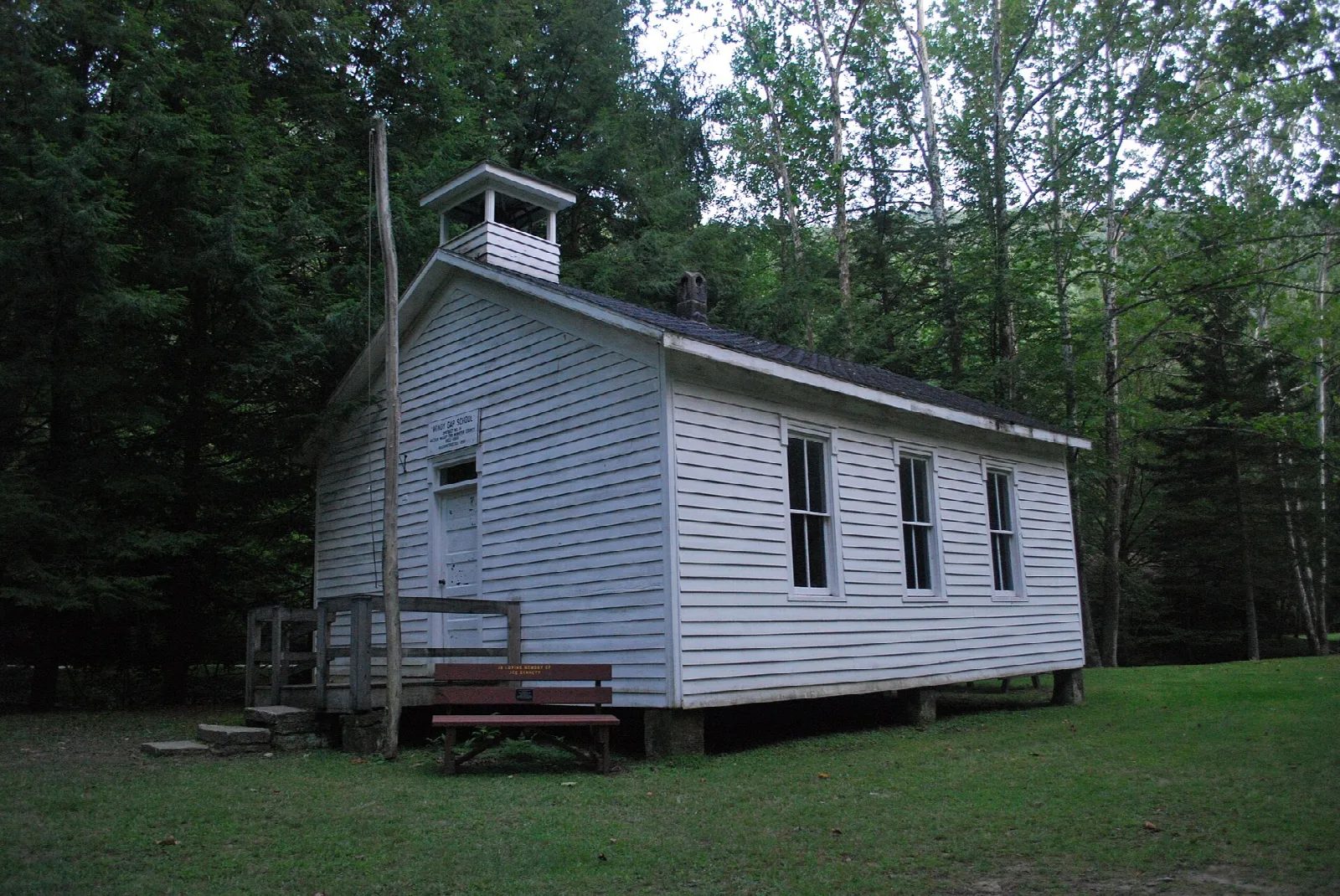 White one-room schoolhouse with a small cupola beside a lawn and dark woods