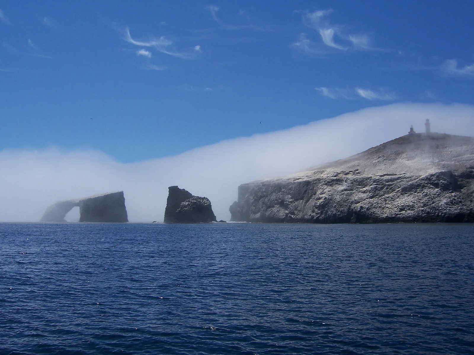 A rock arch and island cliffs rising from deep blue sea, with a bank of fog drifting behind
