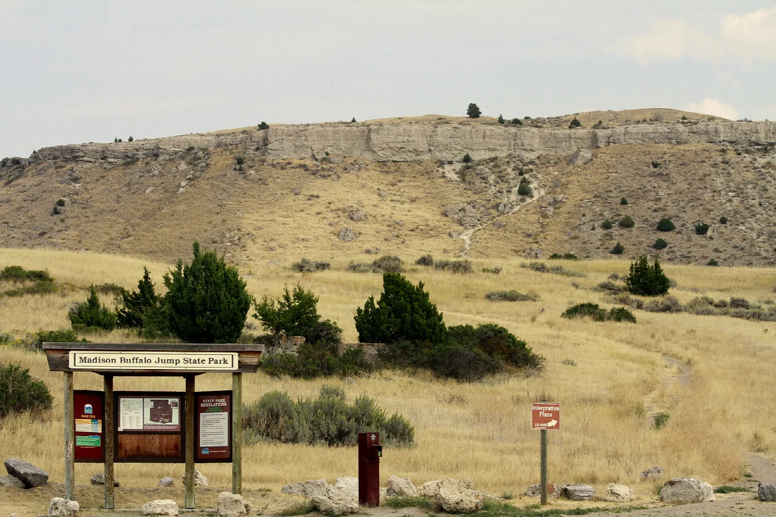Grassy hillside below a long limestone cliff, with an information kiosk in front