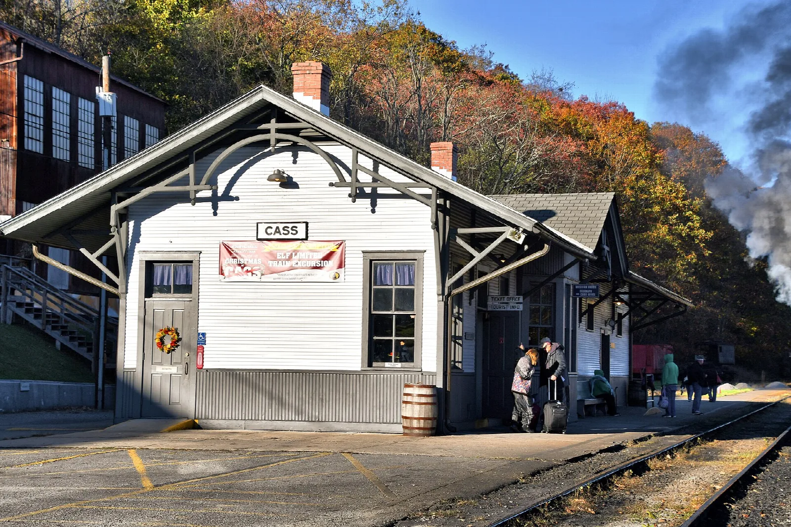 White depot with a ticket window and steam rising beside the tracks under fall trees