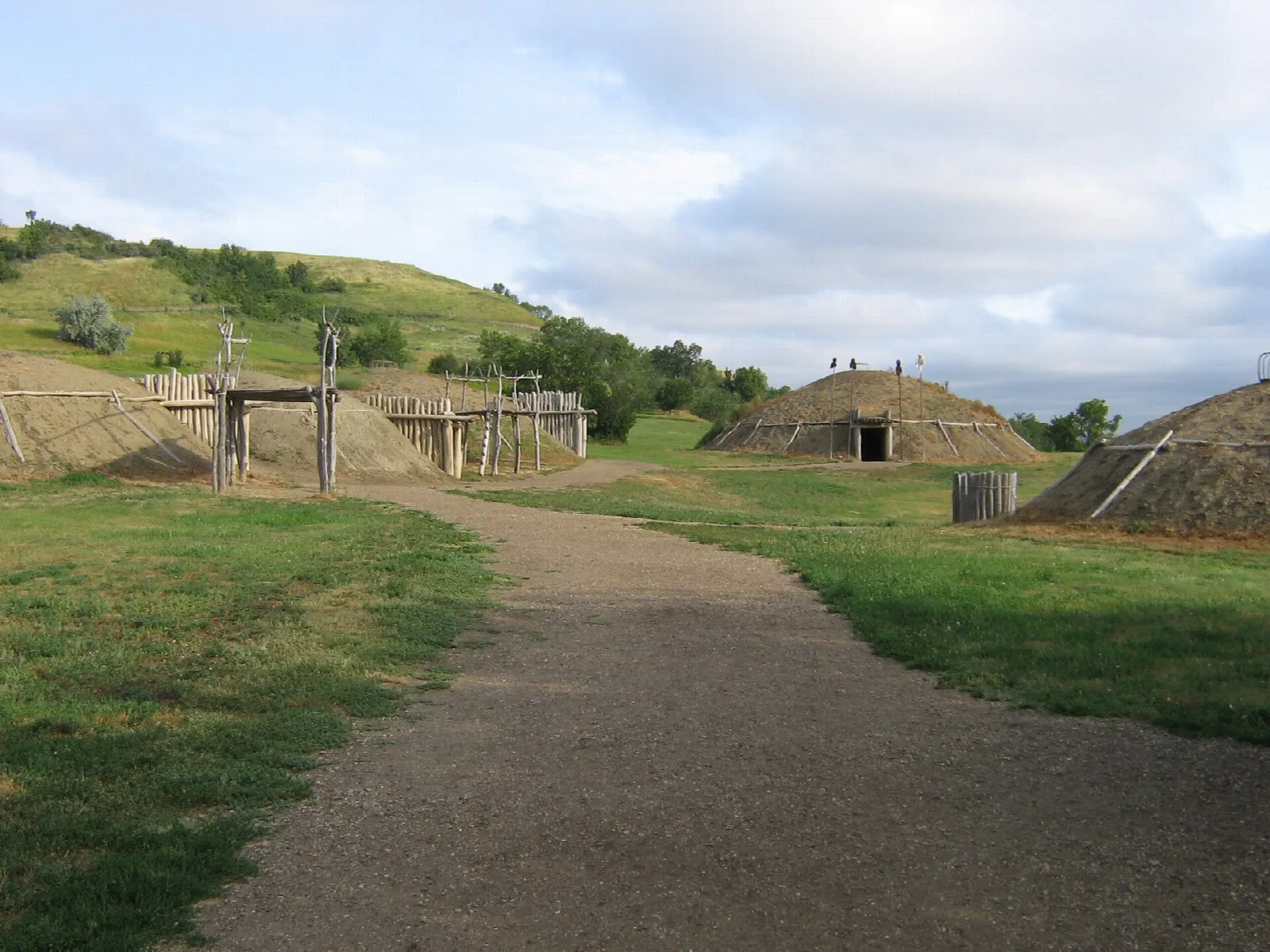 Gravel path leading toward grass-covered earthlodges on a hillside under a cloudy sky