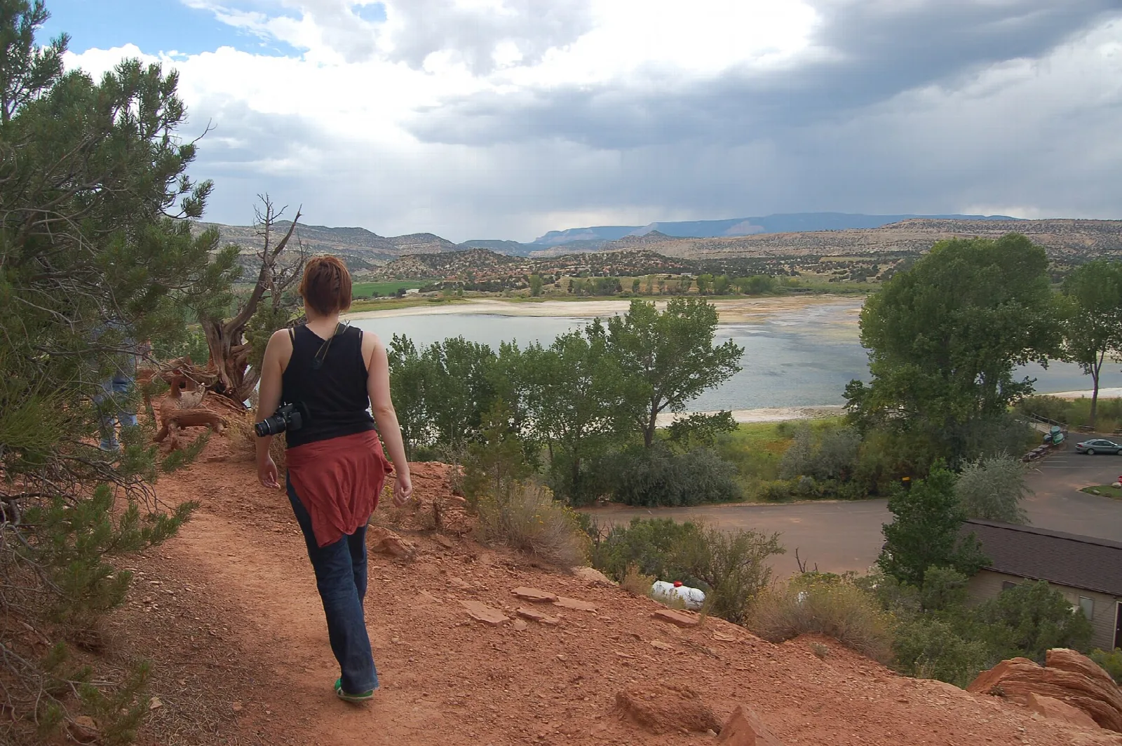 Hiker walking a red dirt trail above a reservoir under stormy sky