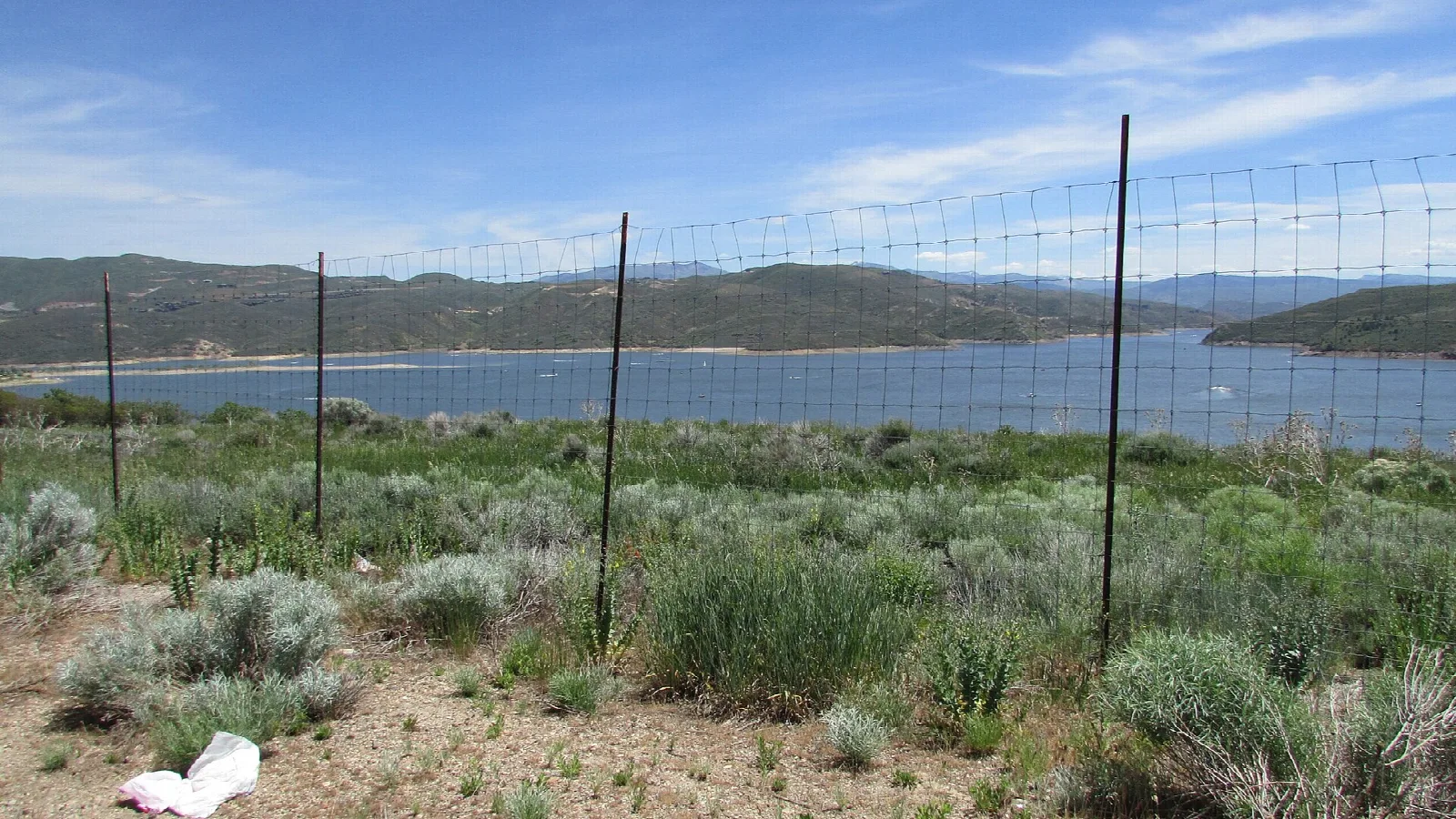 Sagebrush hillside with a fence above a reservoir and green hills beyond