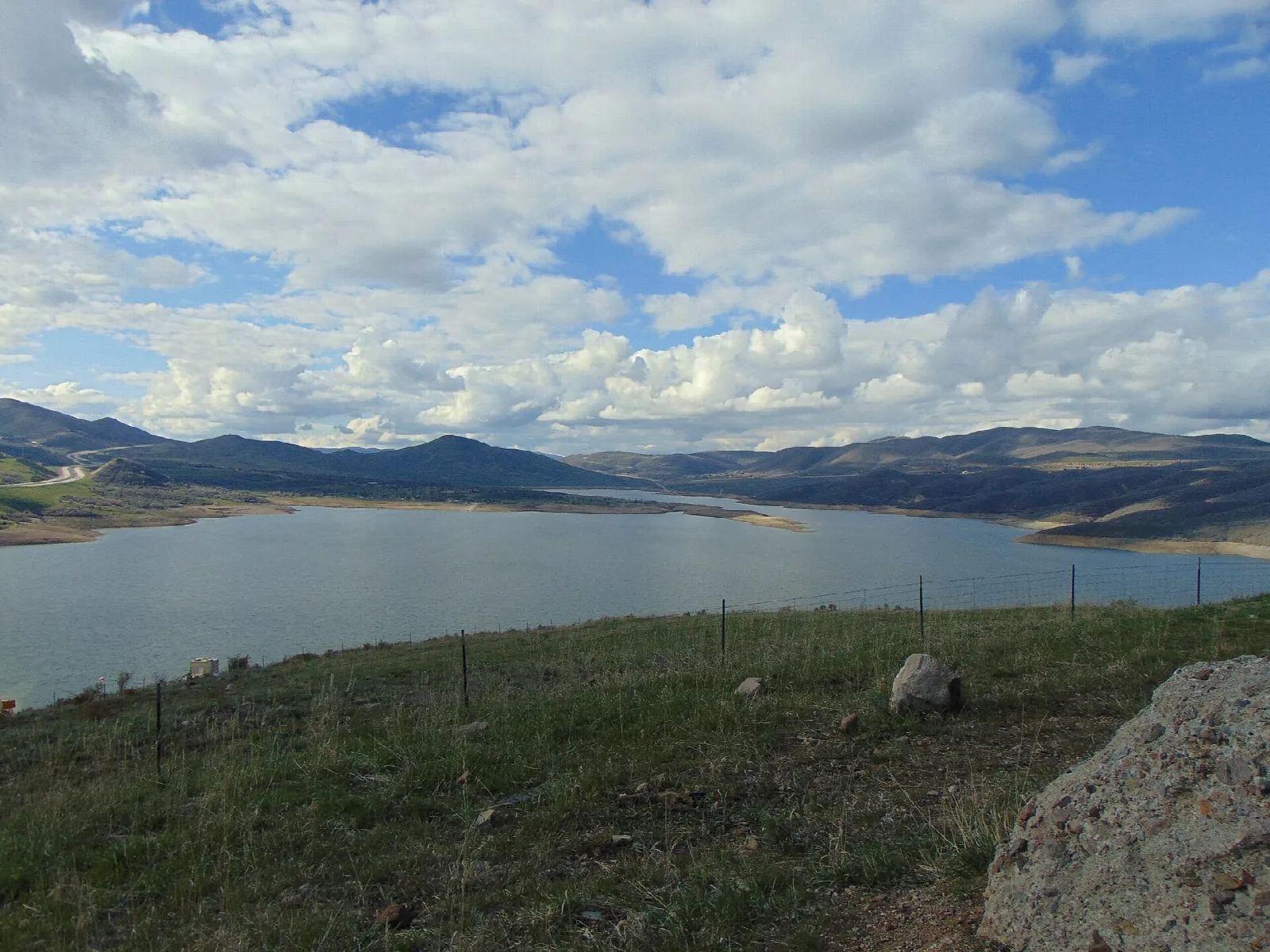 Wide blue reservoir below grassy hills and mountains under a cloudy sky