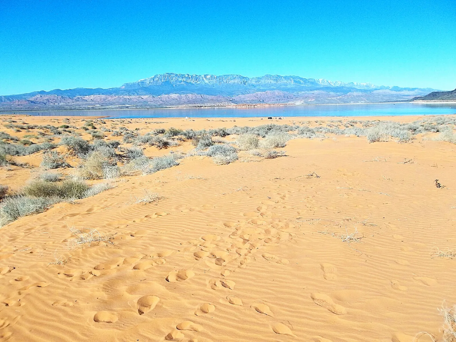 Rippled orange sand dunes beside a reservoir with distant mountains under a bright blue sky
