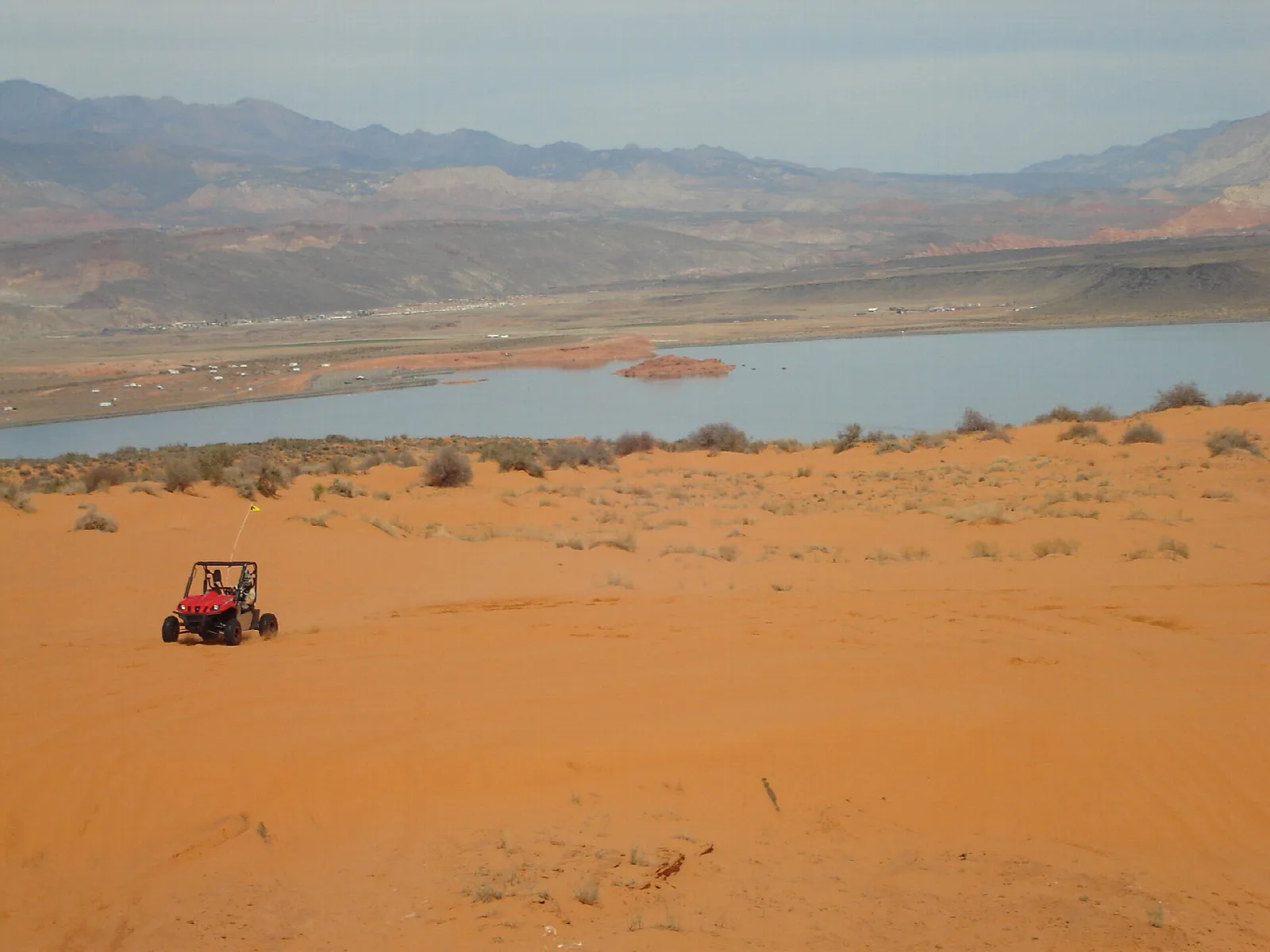 A red off-road vehicle on orange sand above a reservoir with mountains beyond