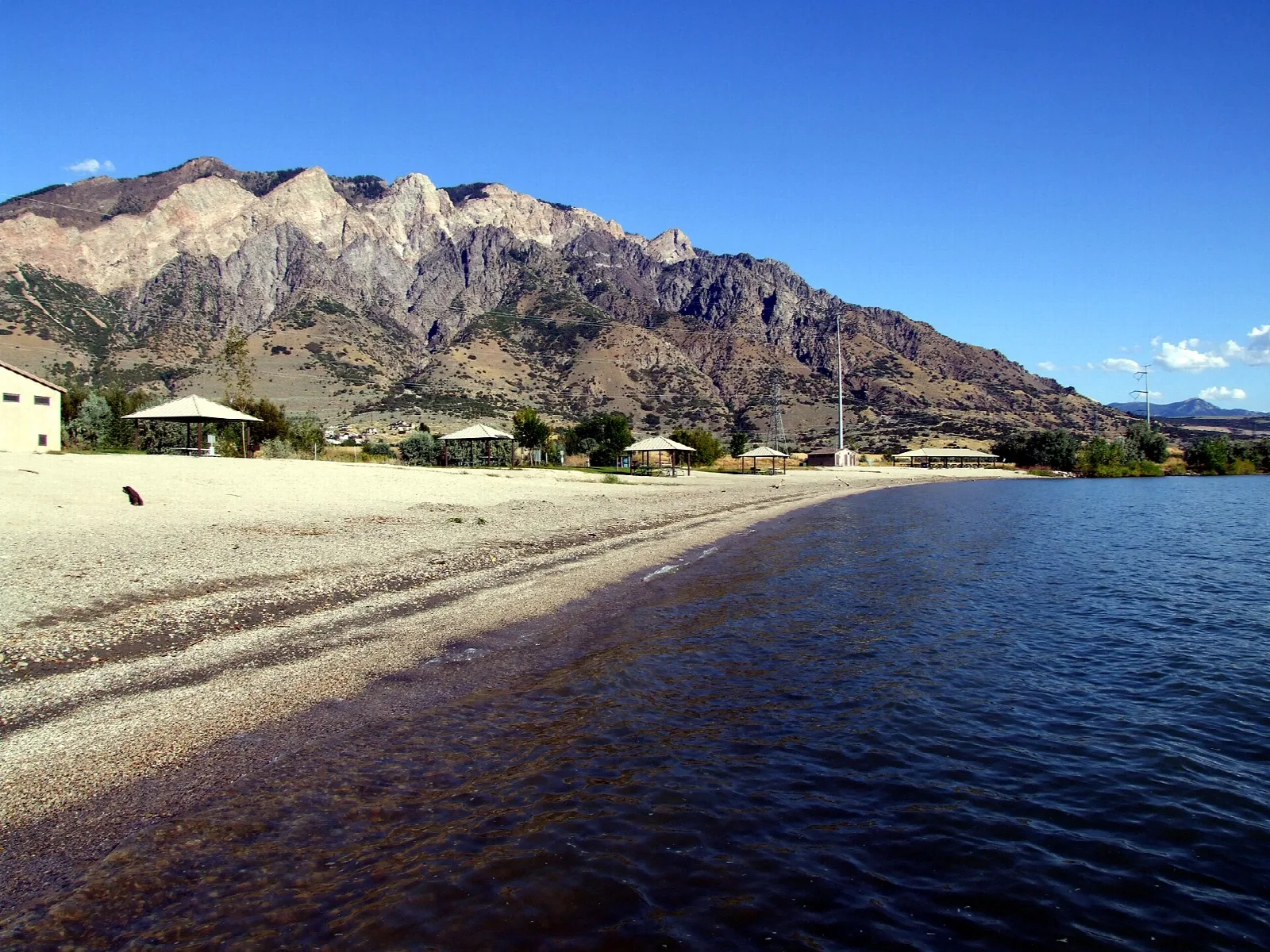 Sandy beach and dark water with a rugged mountain range behind under a blue sky