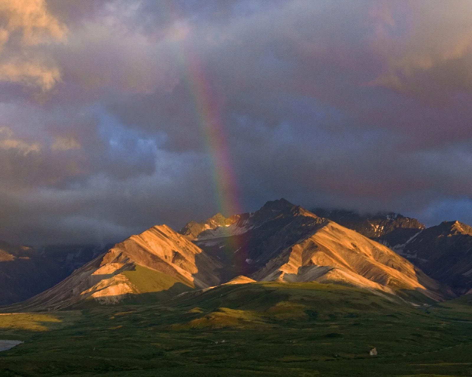 A rainbow over sunlit, multicolored mountains under dark storm clouds