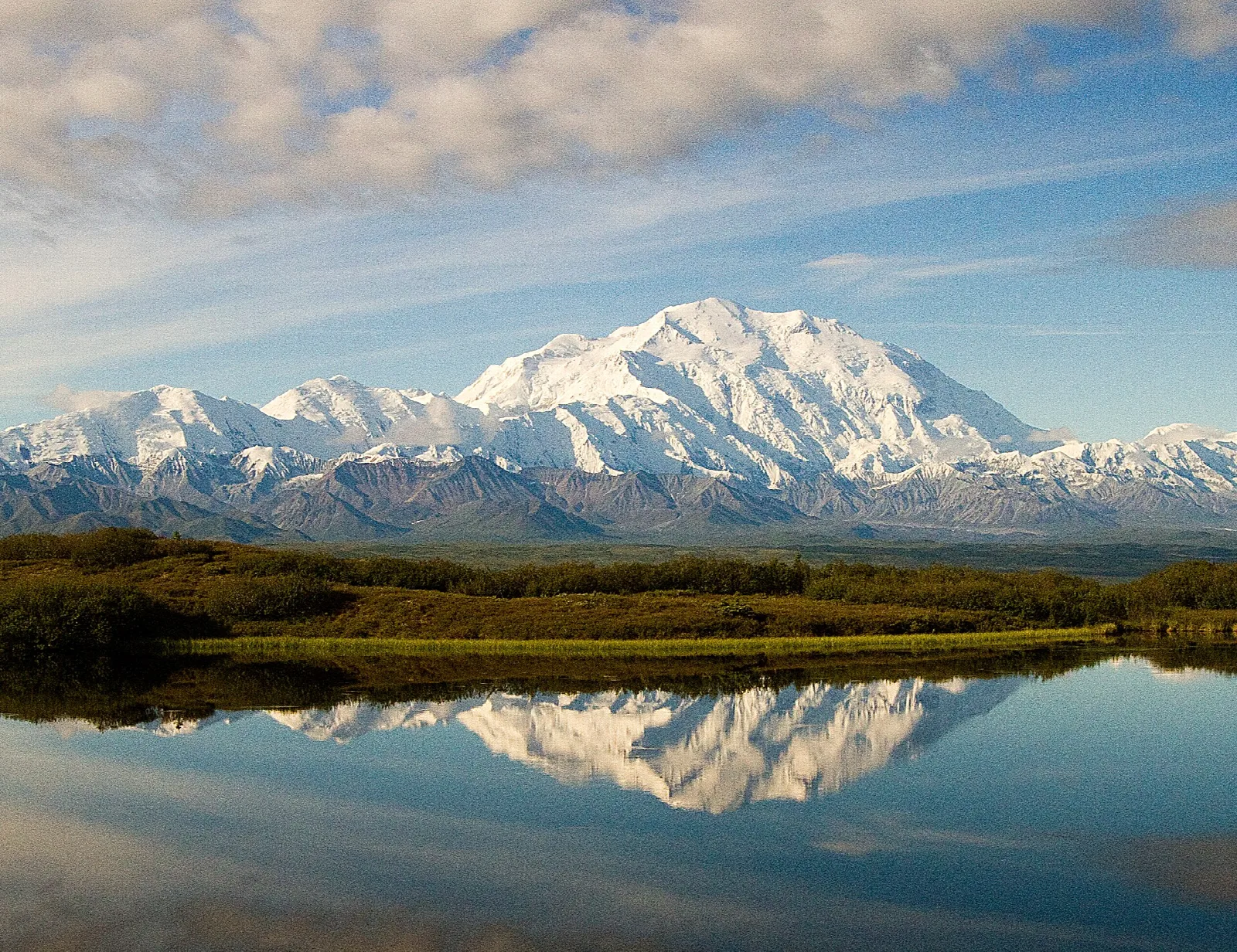 A massive snow-covered mountain reflected in a calm lake under a blue sky