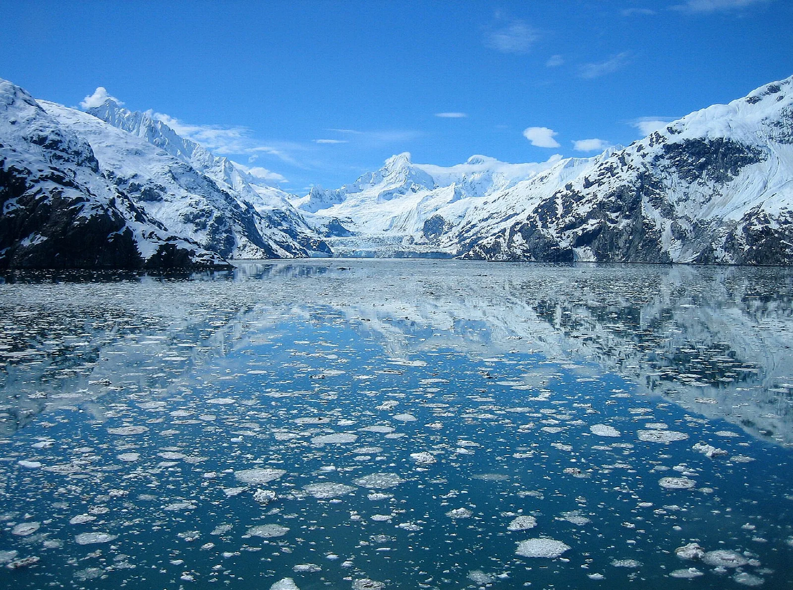 Ice-dotted water reflecting snowy peaks and a tidewater glacier under a blue sky