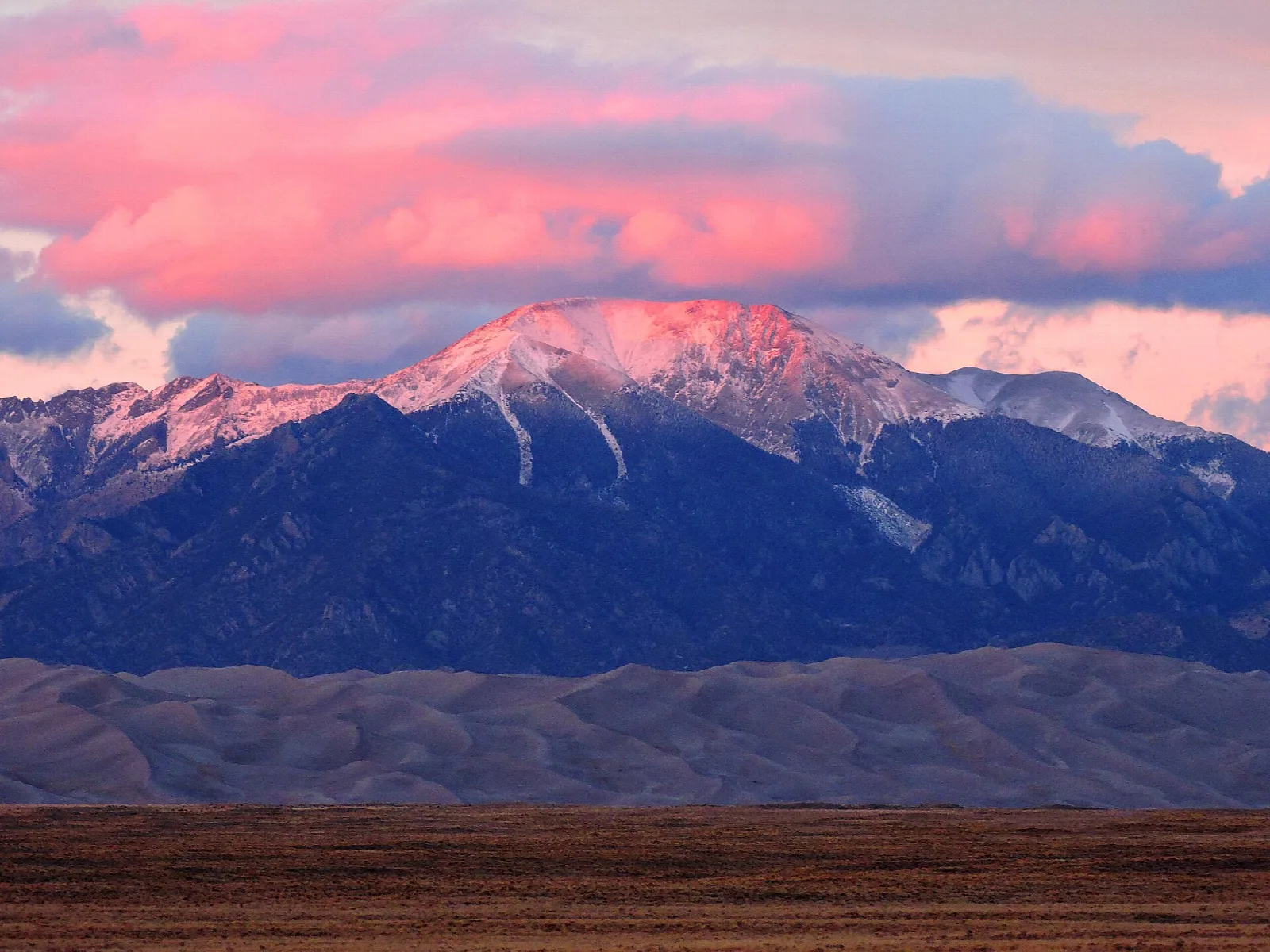 Pink evening light on a snowy peak rising above the dunefield