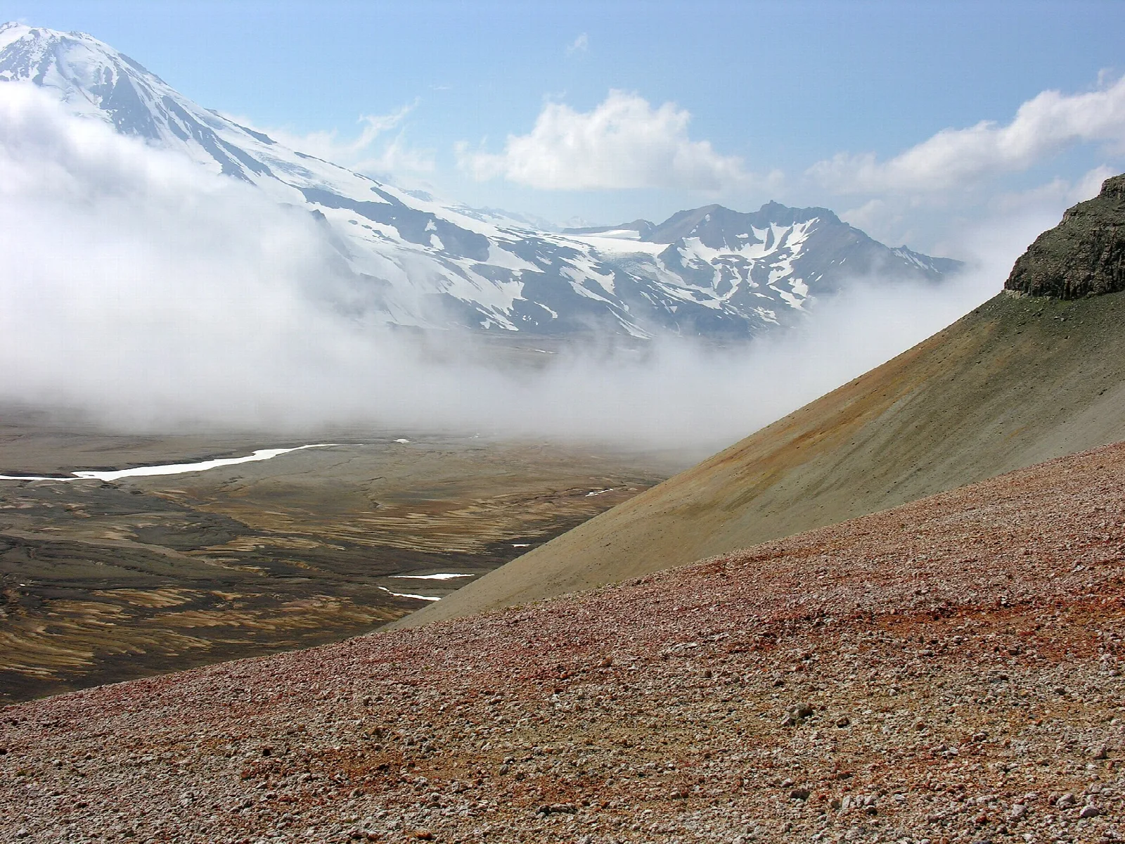 Colorful ash slopes and a deep valley below clouded, snowy volcanoes
