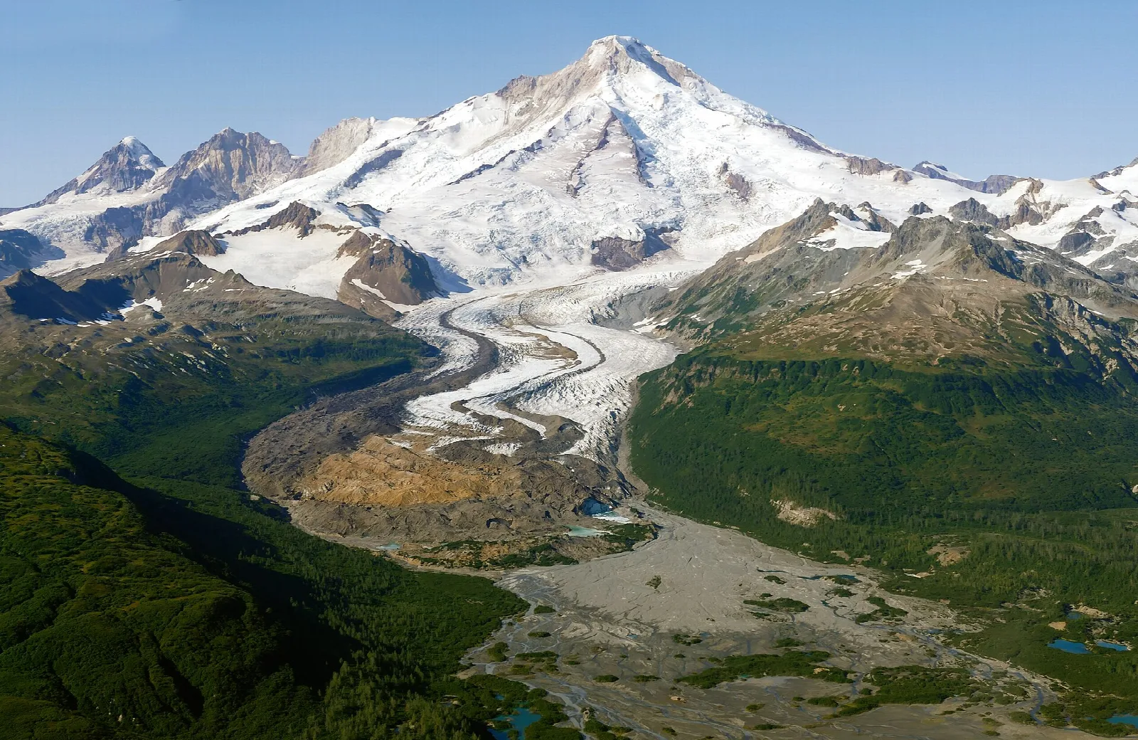 A glacier flowing from a snowy volcano down to a green, braided river valley