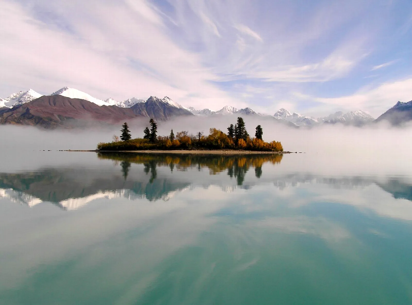 A small island of autumn trees in a still lake, fog drifting below pink-lit mountains