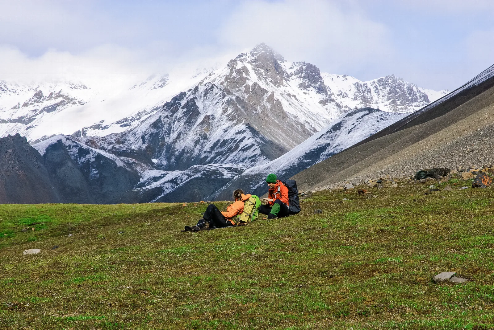 Two backpackers sitting in an alpine meadow with snowy mountains in the background