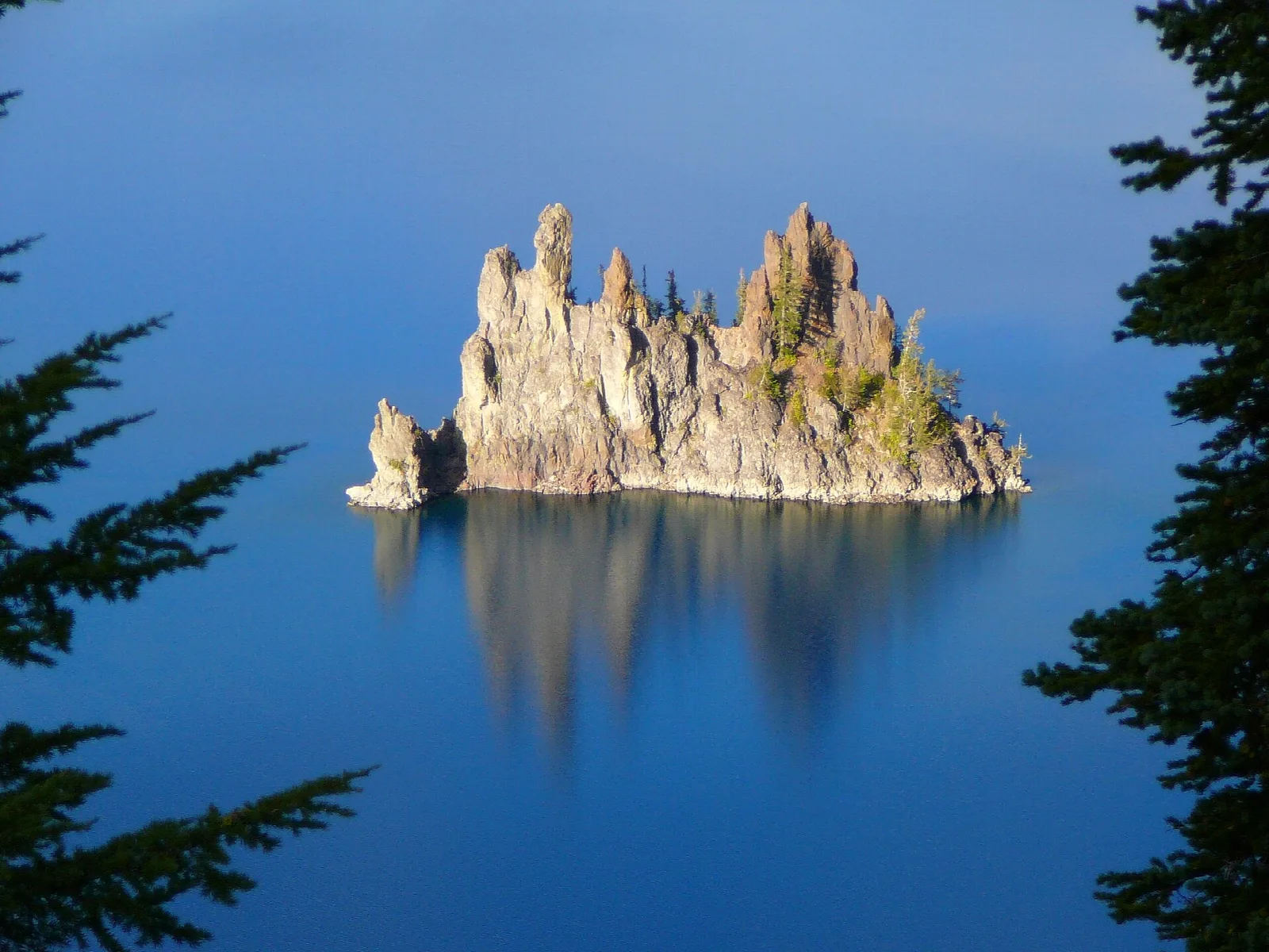 A jagged rock island in calm, deep blue water, framed by evergreens