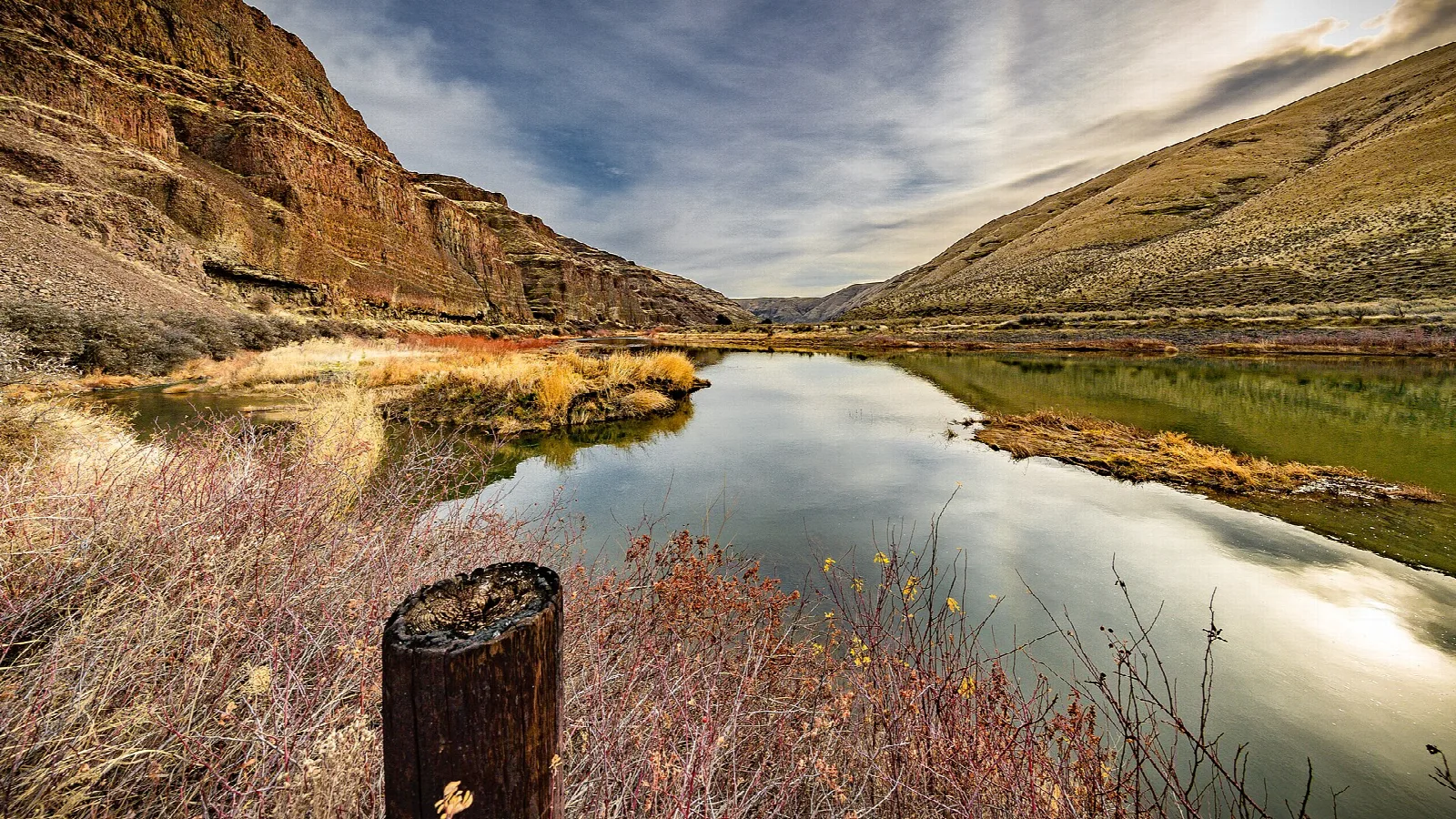 Calm river reflecting canyon walls and clouds, with a post in the foreground