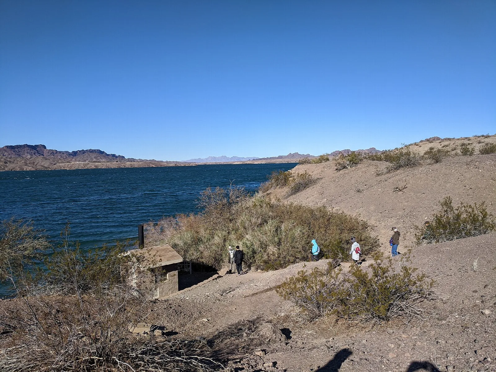 Hikers on a sandy shoreline path above the blue lake