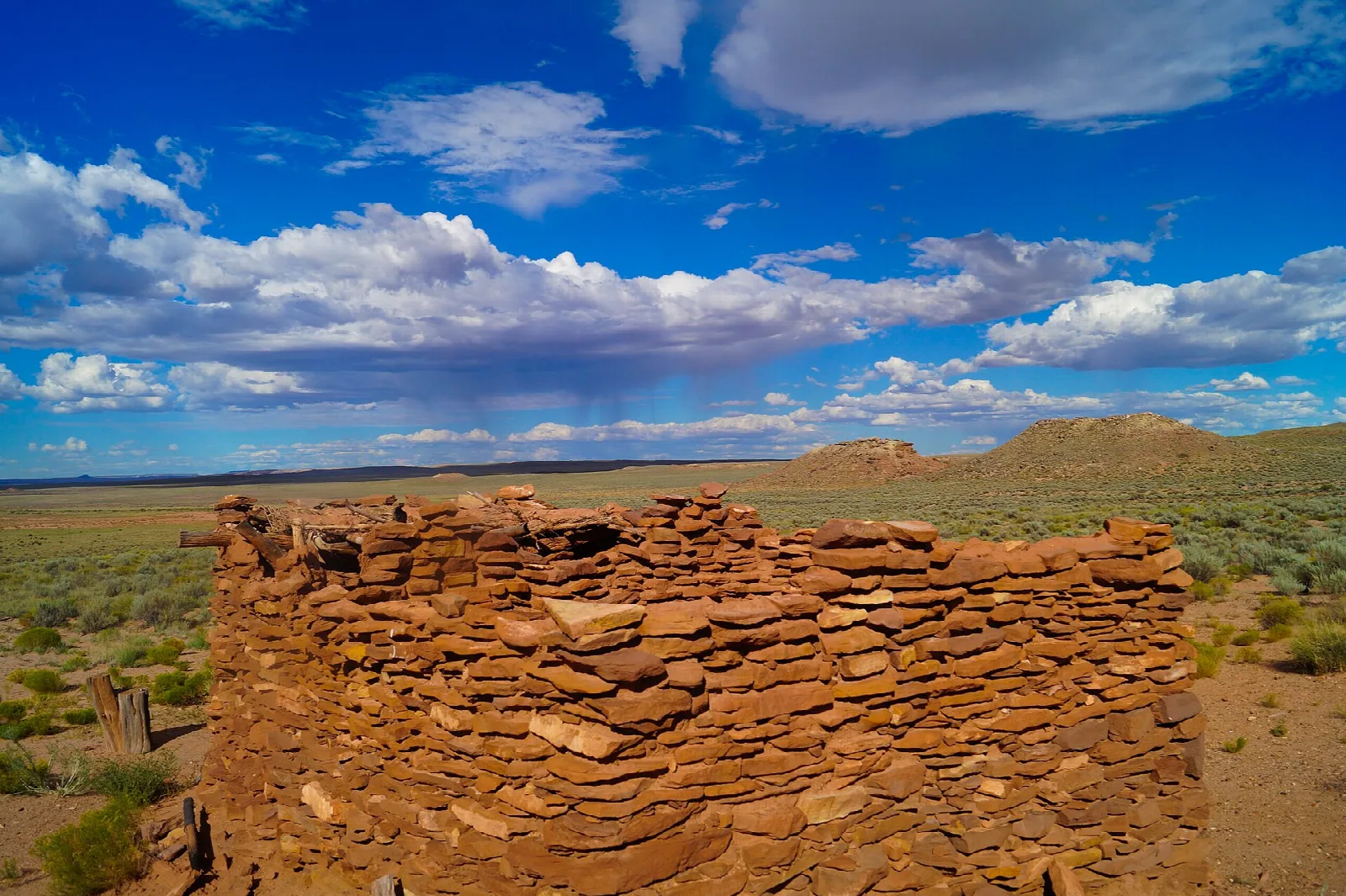Low stacked-stone pueblo walls on open grassland under blue sky