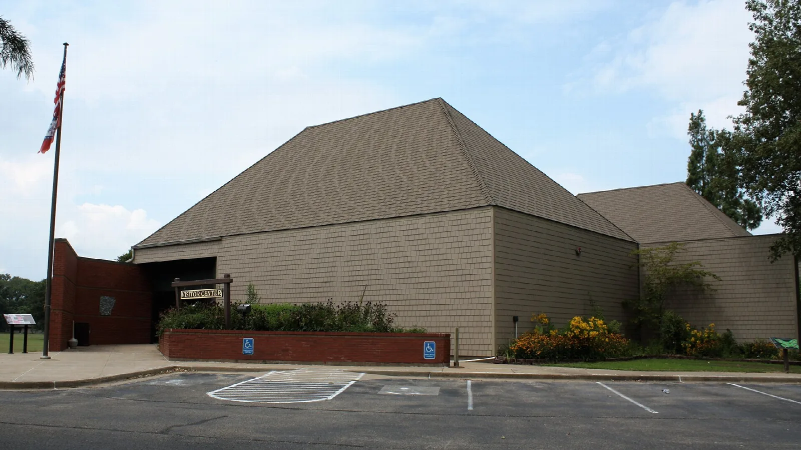 Low tan museum building with a flag and a parking lot in front