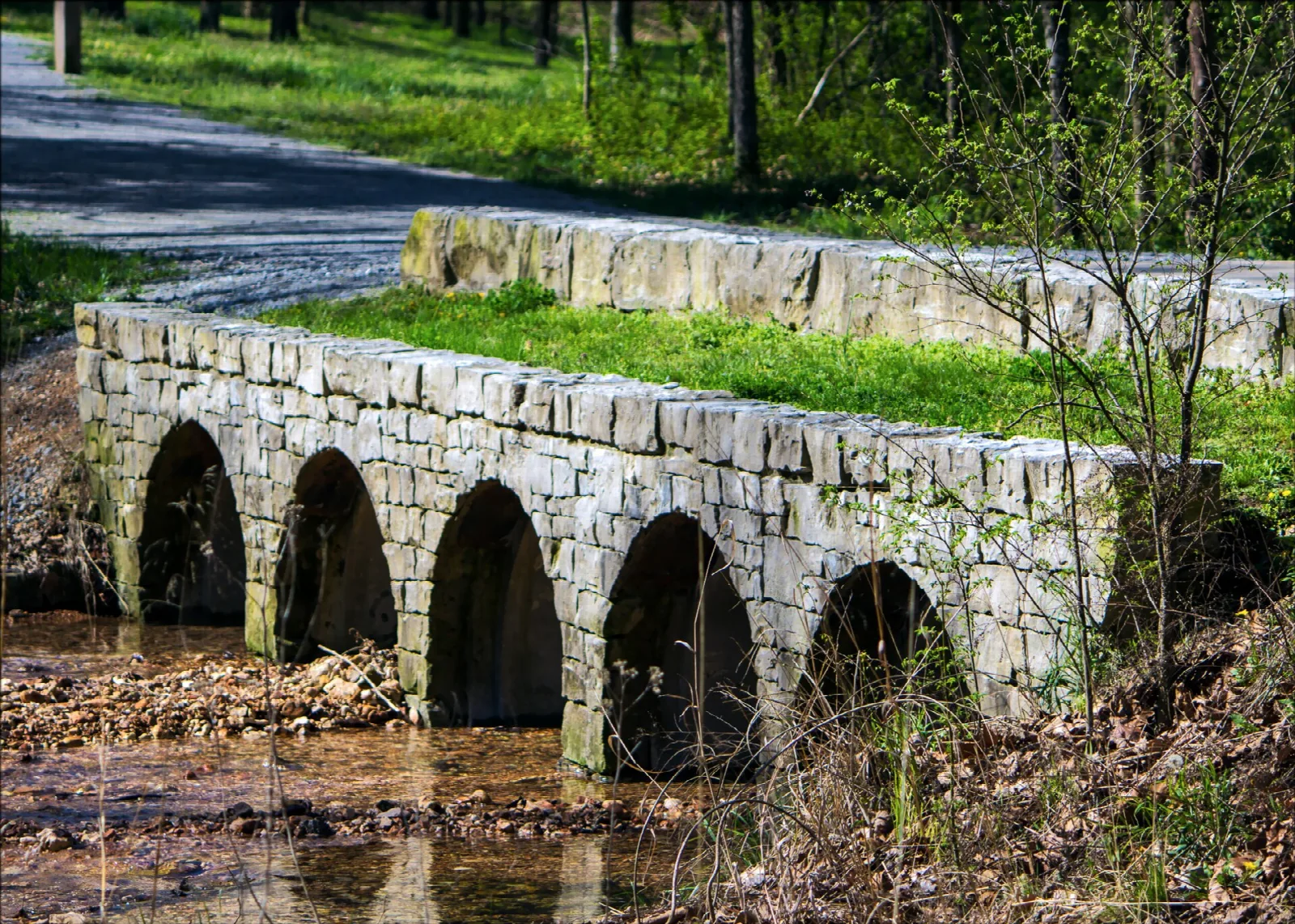 Arched stone bridge over a creek beside a road, with green grass above