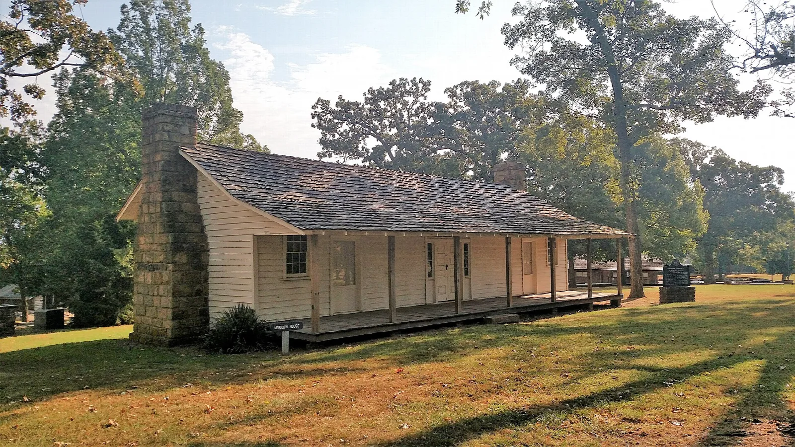 Single-story historic house with a porch and stone chimney under big trees